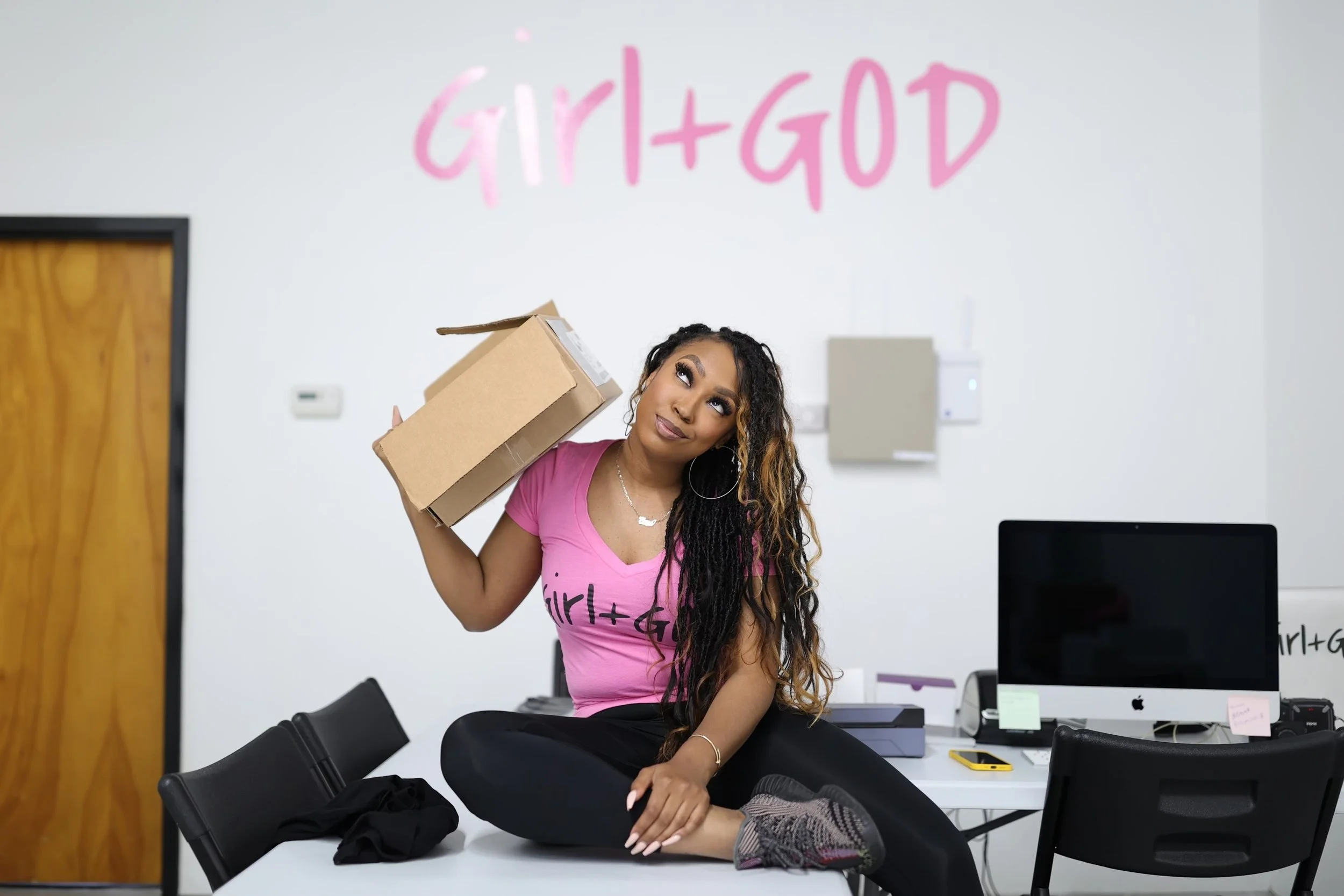 A young woman with long curly hair and hoop earrings, wearing a pink t-shirt, sitting at a desk in an office, holding a cardboard box on her shoulder, looking upwards with a thoughtful expression.