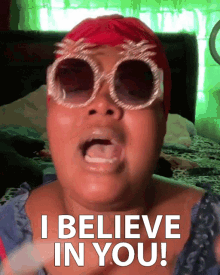 Person wearing large sunglasses with a red wig, shouting with an emotional expression, with the caption 'I BELIEVE IN YOU!'