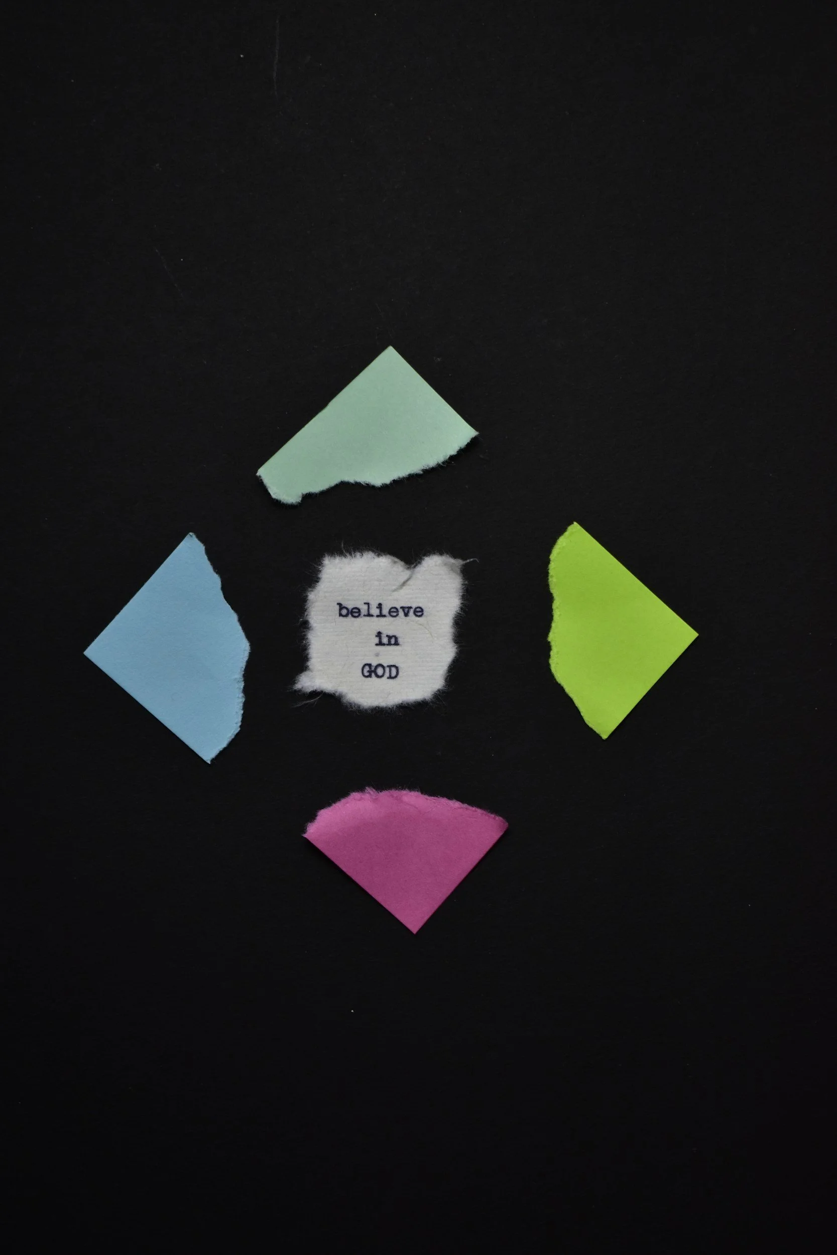 A black background with torn paper pieces in various colors, including blue, green, pink, and white, with the phrase 'believe in GOD' written on a torn white piece of paper.