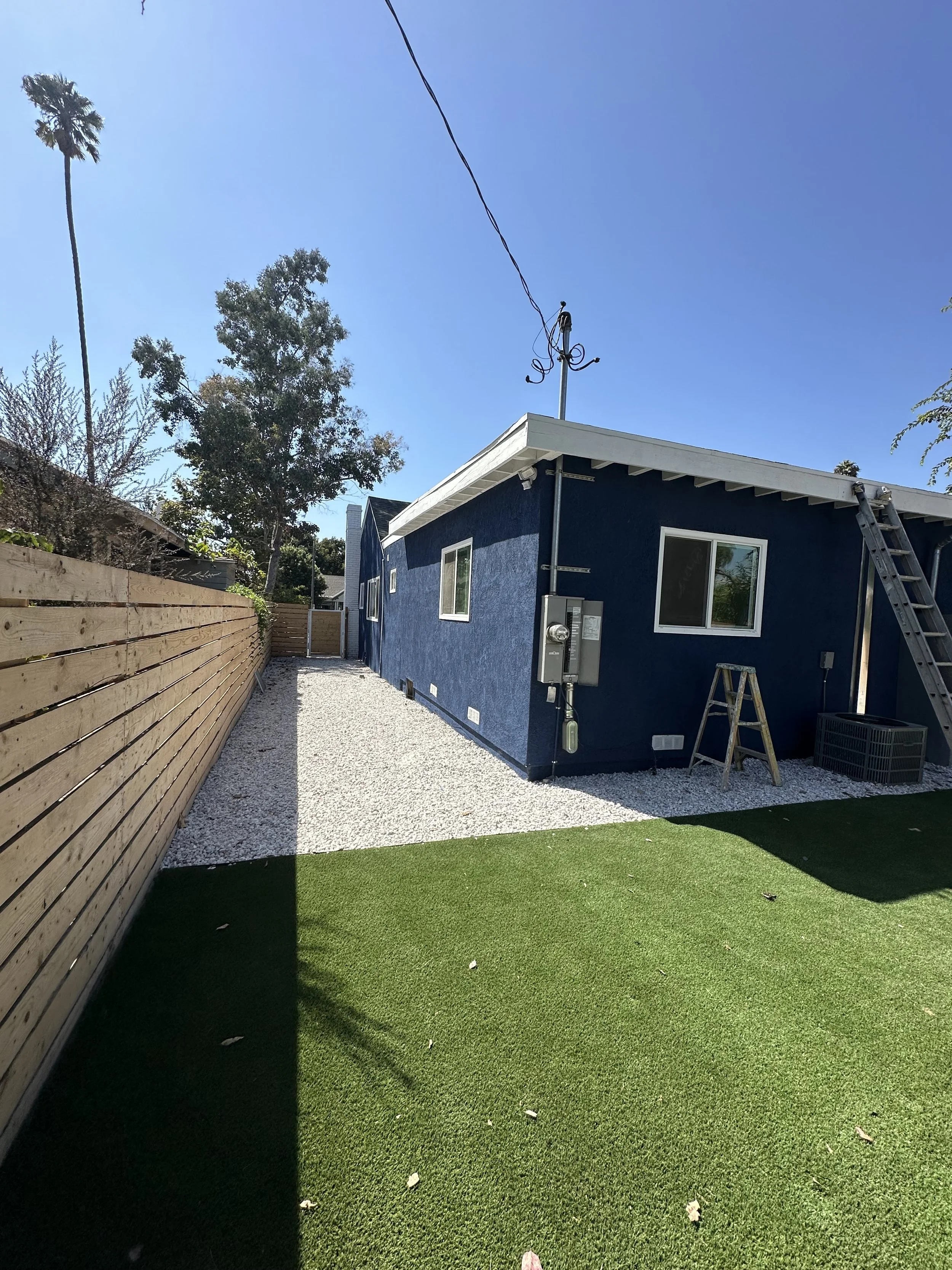 A backyard with a small patch of green artificial grass, a blue house with white trim, a wooden fence on the left, and trees in the background. A ladder leans against the house, and the sky is clear and sunny.