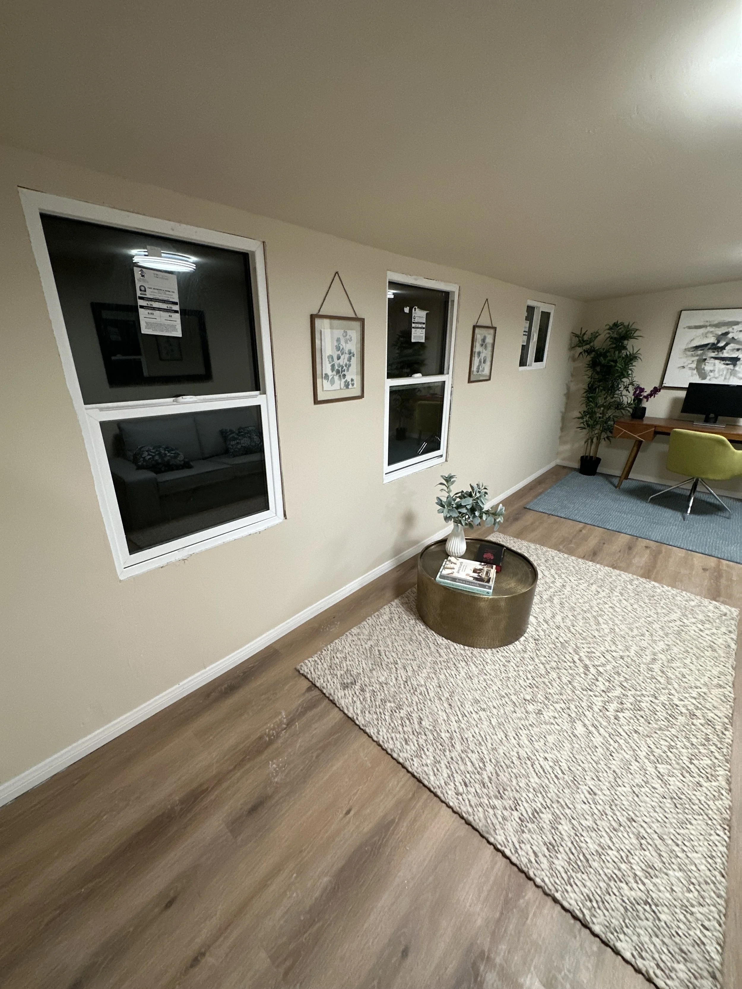 Living room with beige walls, three windows, framed artwork, a potted plant, a desk with a monitor, a yellow chair, a blue rug, and a round coffee table with magazines.