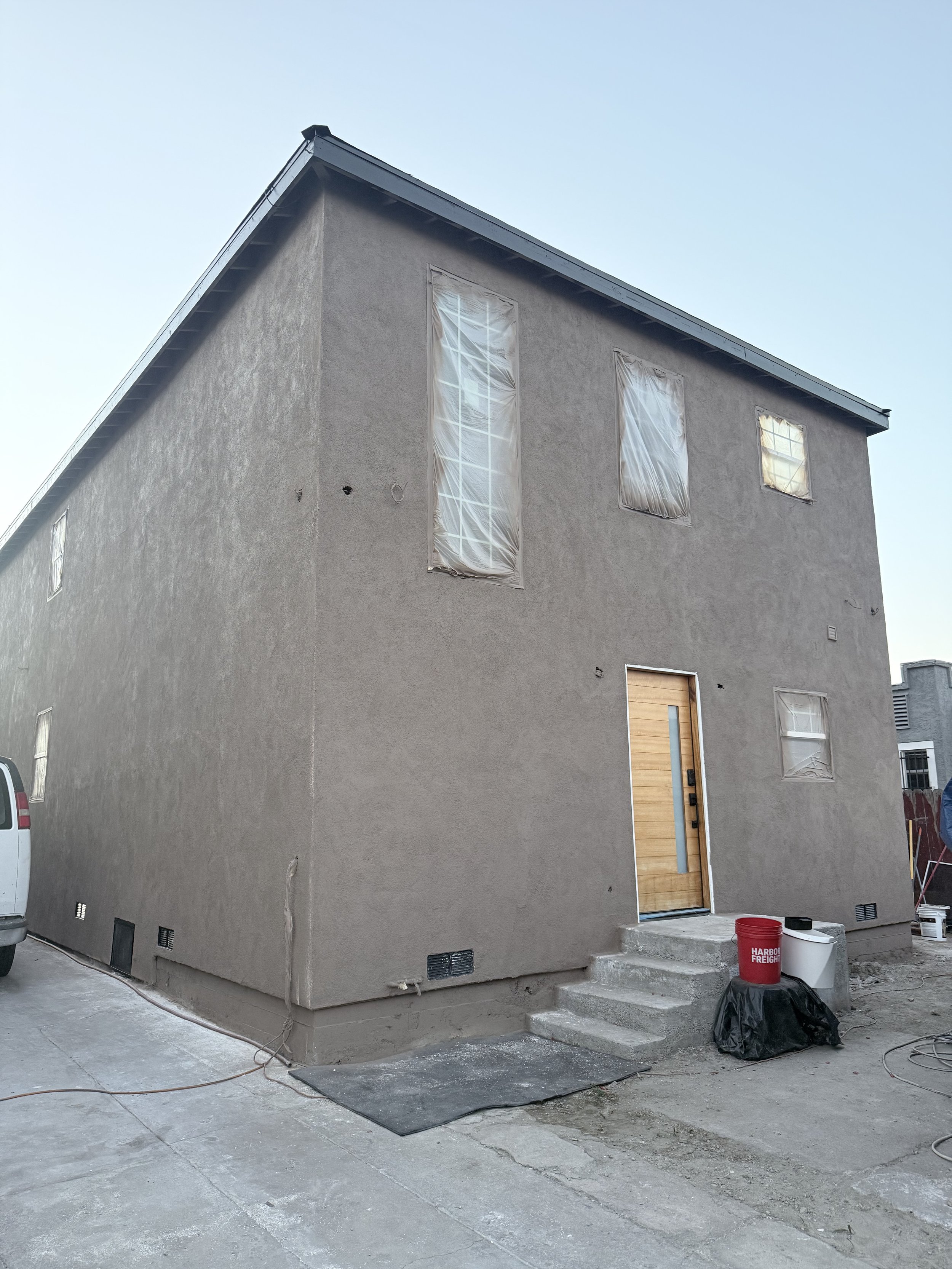 Front of a house under construction with a concrete facade, a wooden front door, covered windows, and tools and supplies on the ground.
