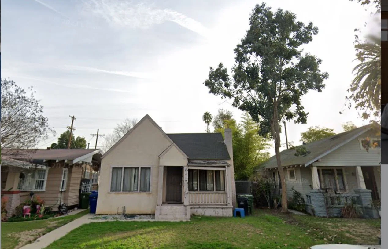 An old, small, white house with a front porch and a steep roof, surrounded by trees and neighboring houses, under a clear sky.