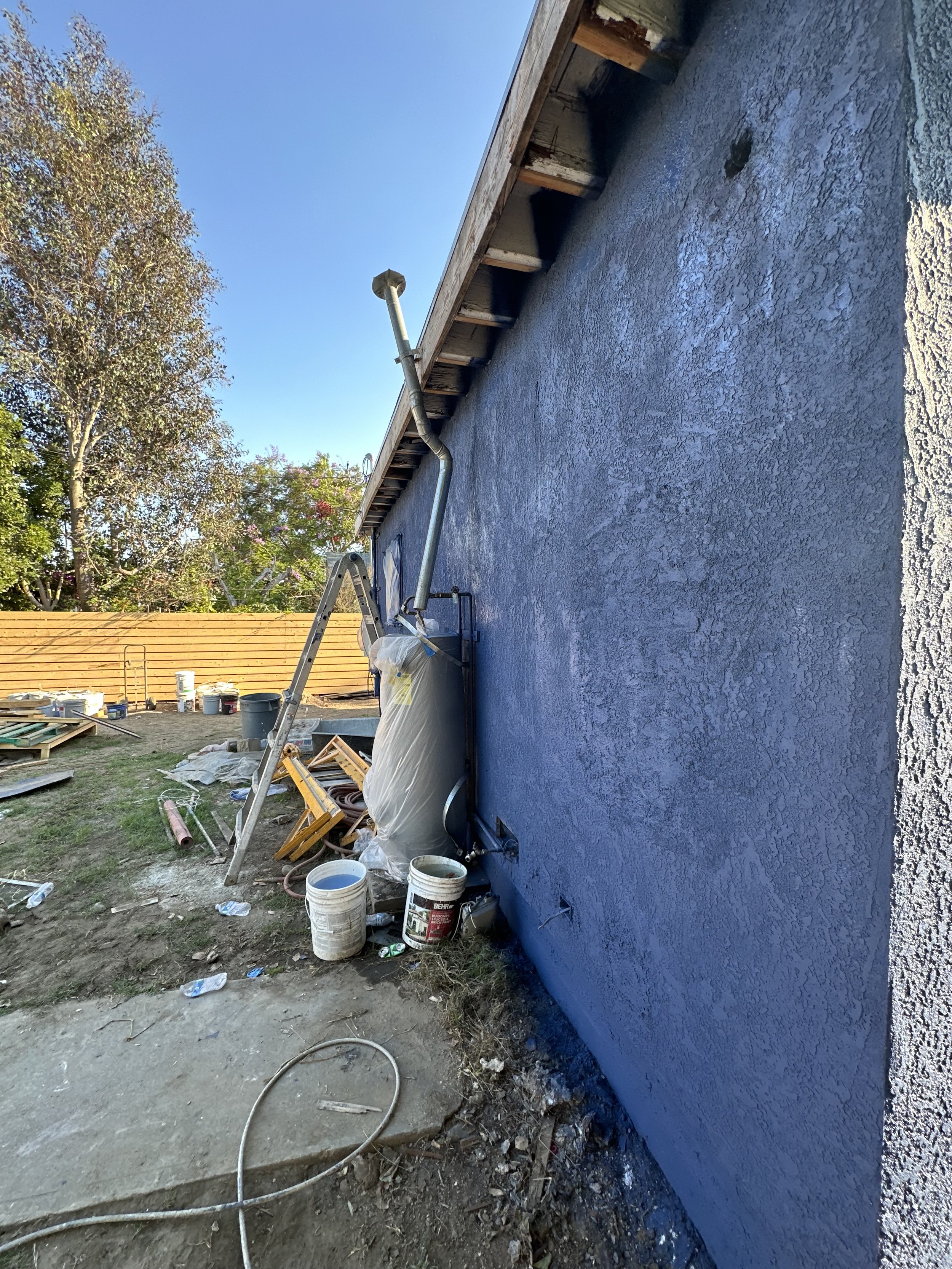 Construction site with a partially painted blue wall, ladders, buckets, and construction materials on the ground, with trees and a wooden fence in the background.