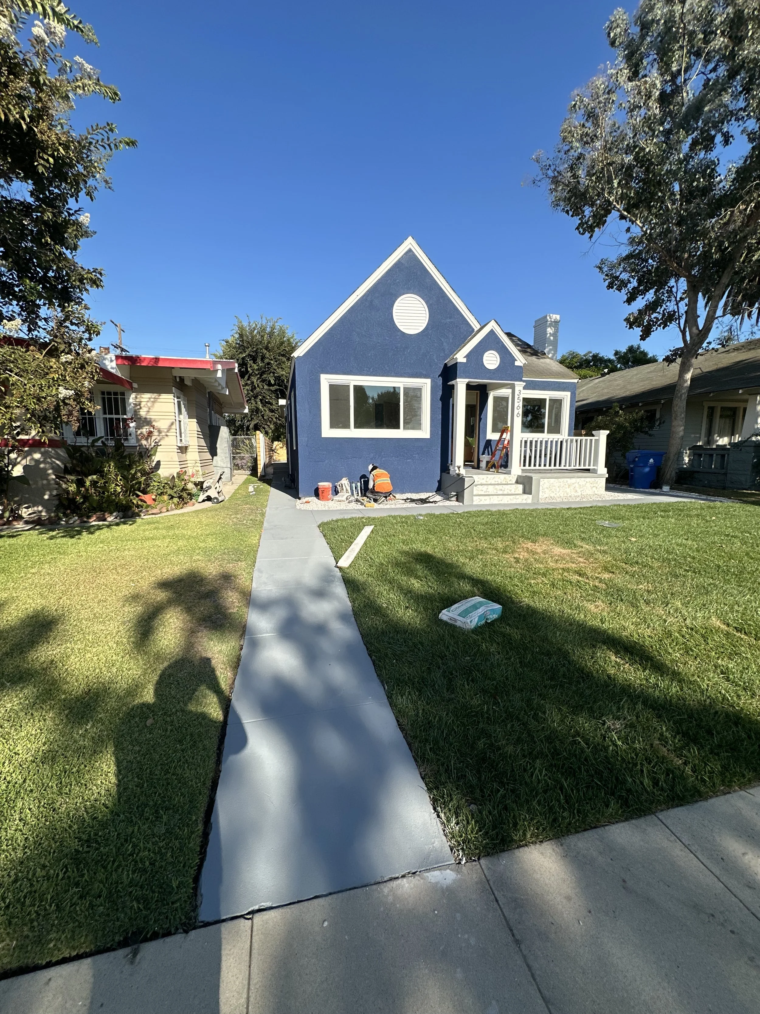 A small blue house with white trim in a residential neighborhood, with a sidewalk and a lawn in the foreground, and another house and trees in the background. There is a worker in an orange safety vest and hat working near the front steps of the house.