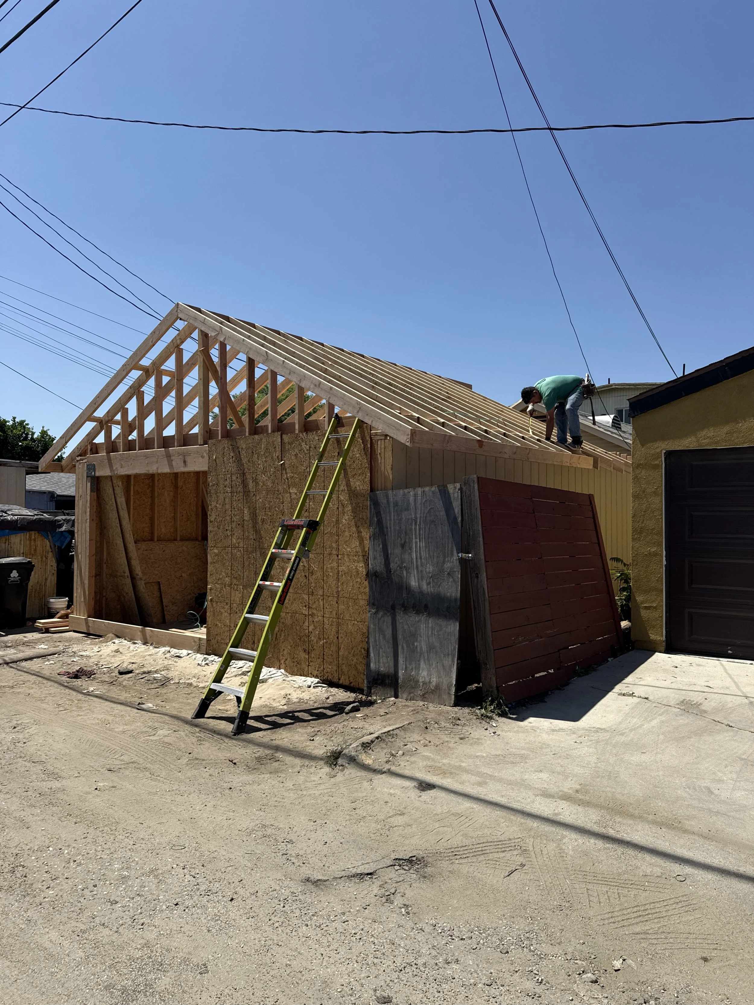 Construction site of a house with wooden framing on the roof, a person working on the roof, a yellow ladder leaning against the structure, and a partially built wall with plywood and sheathing. The adjacent building is painted yellow and there is a gravel and dirt street in the foreground.