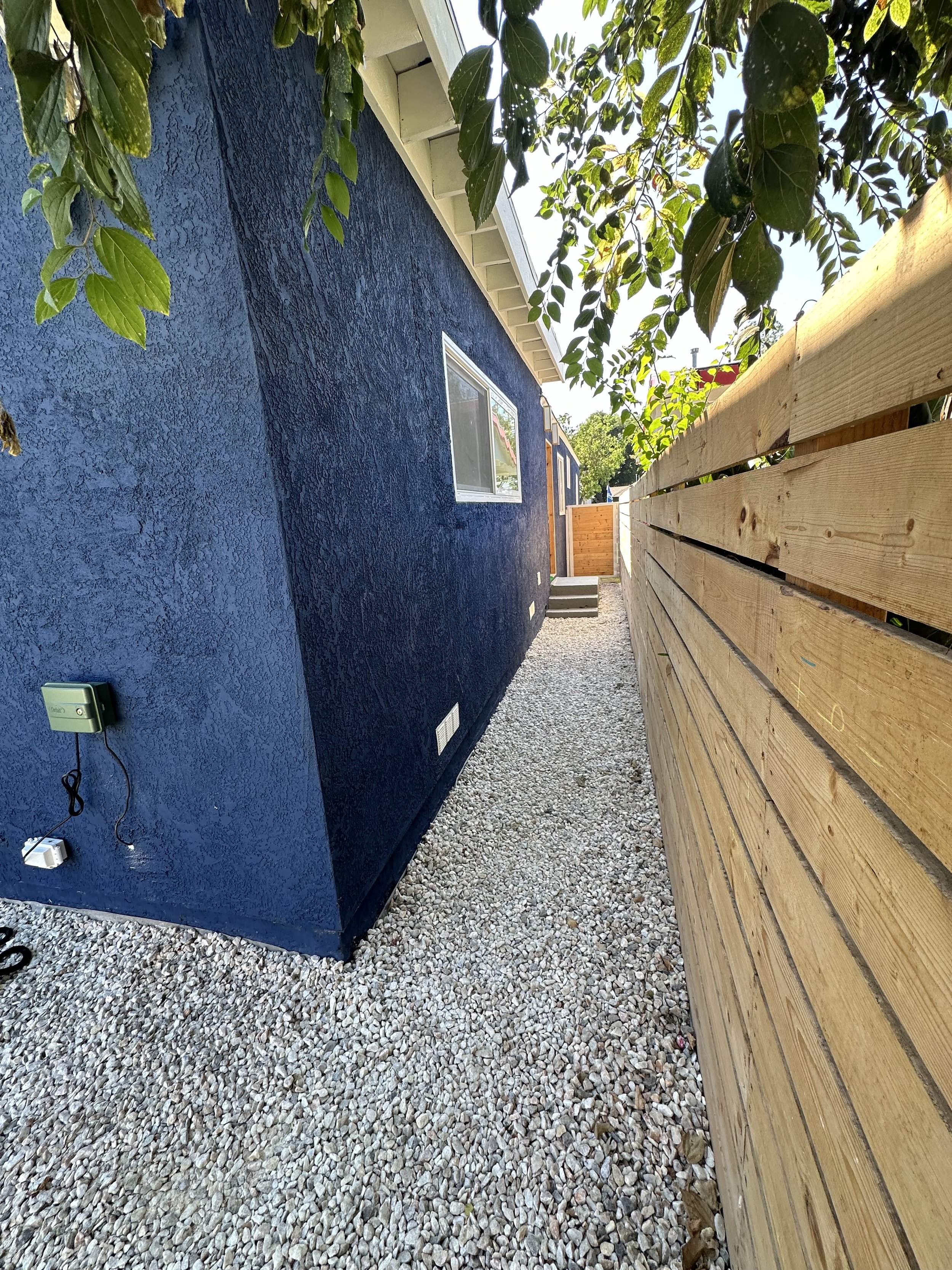 Side view of a house with blue textured exterior wall, window, electrical outlet, and gravel ground, separated from a wooden fence by a small pathway with trees overhead.