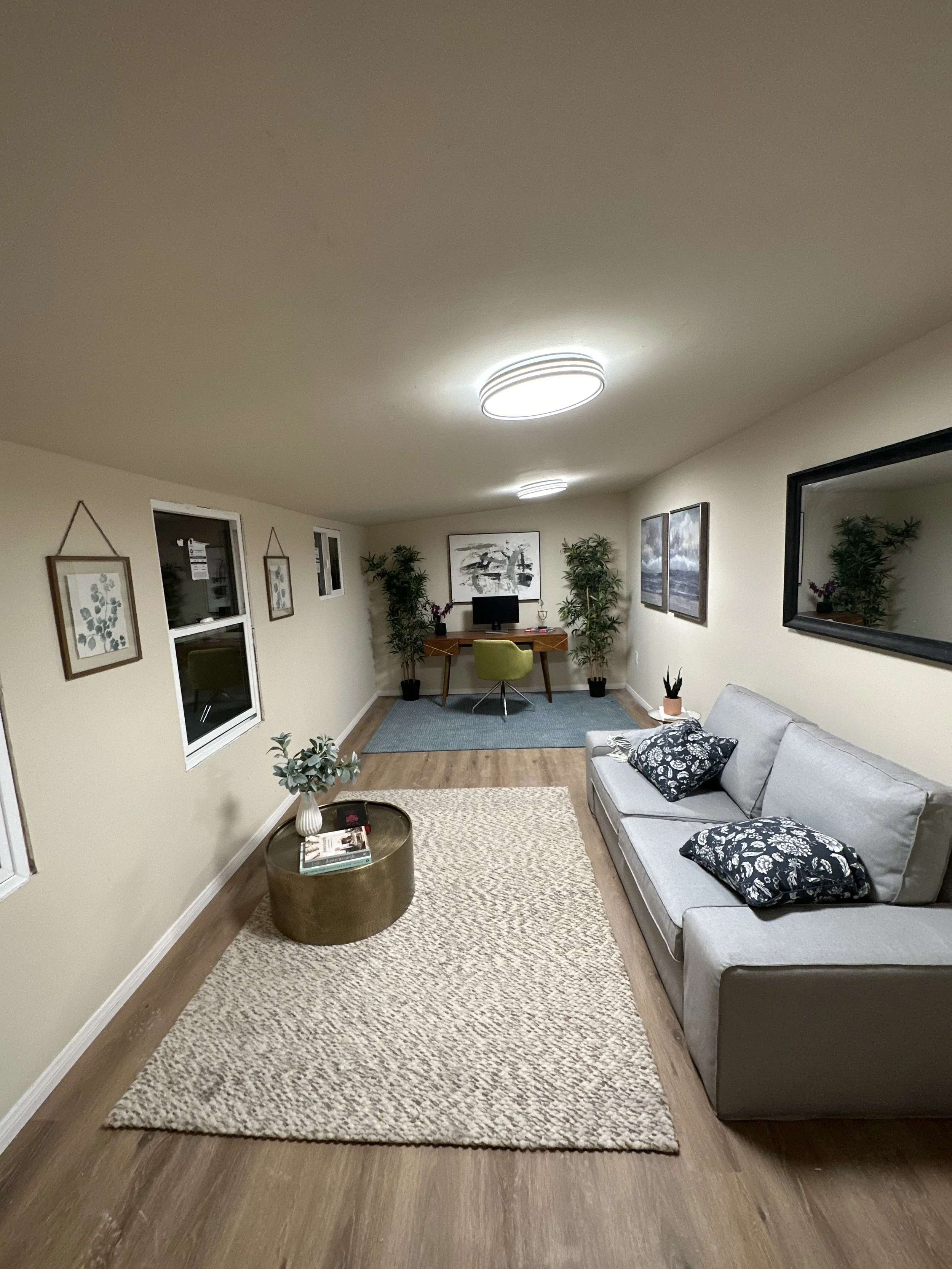 Living room with beige walls, hardwood floors, a light gray sofa with black and white pillows, a shag rug, a small round coffee table with plants and magazines, framed artwork on the walls, a desk with a computer, potted plants, and ceiling lights.