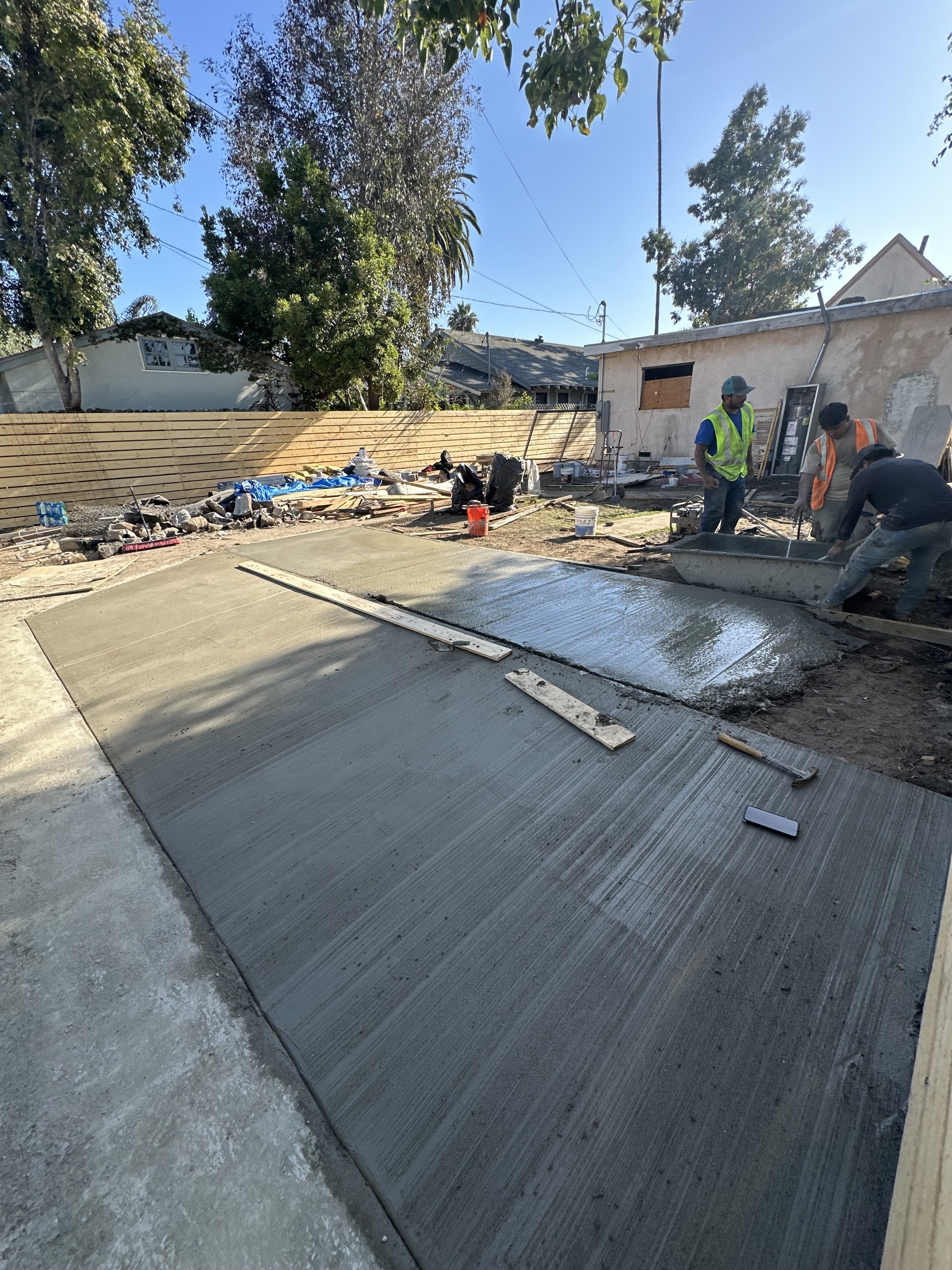Workers pouring concrete on a sidewalk in a residential backyard, with tools and construction materials around.