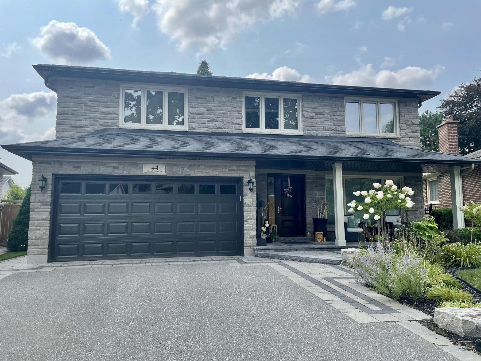 Front view of a modern two-story house with gray stone exterior, black garage door, and large white-framed windows. There is a concrete driveway, a landscaped garden with flowers, and a bright sky with clouds.