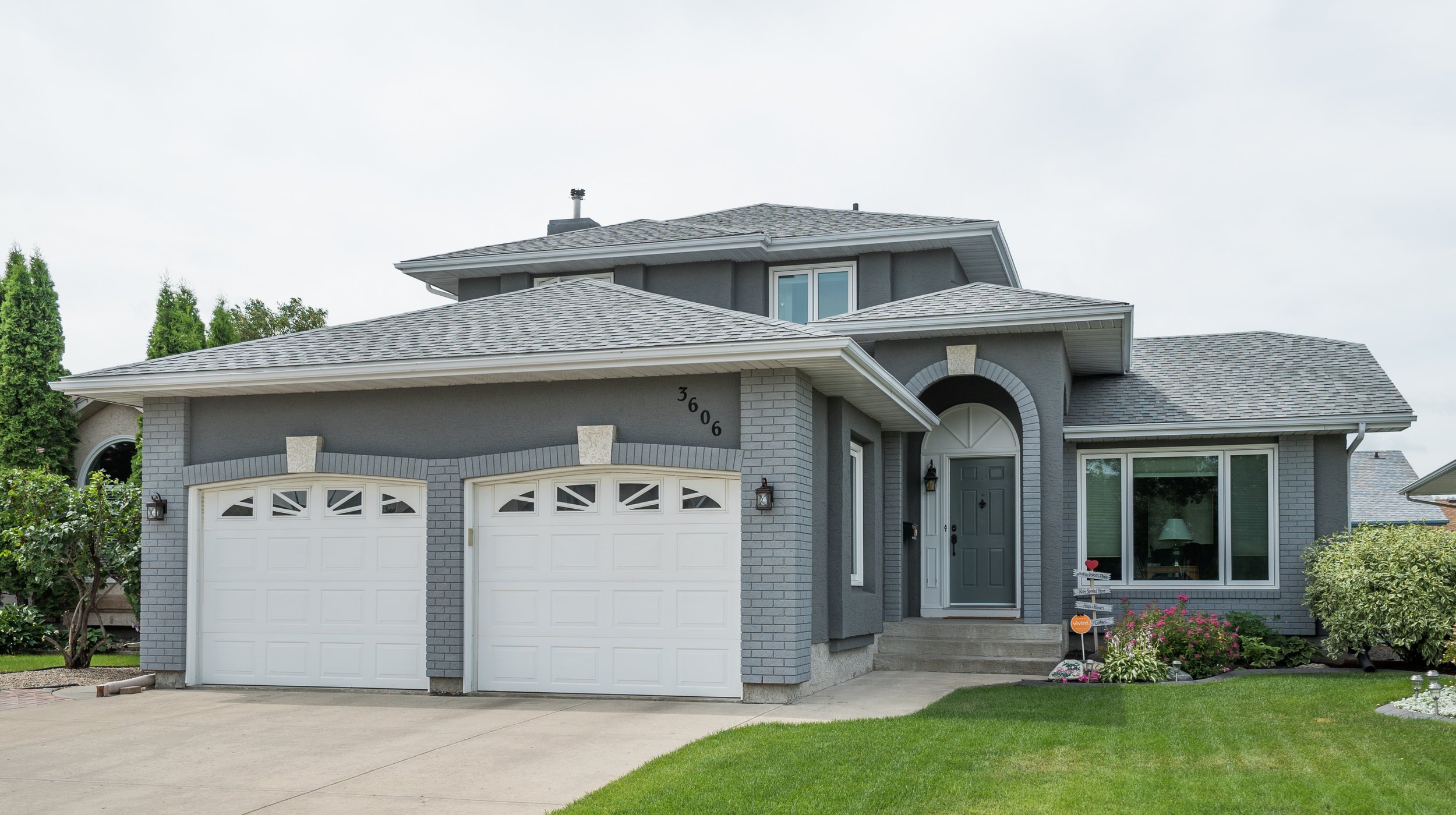 A two-story gray house with white garage doors, a front door, large front window, and landscaping including bushes, flowers, and a lawn.