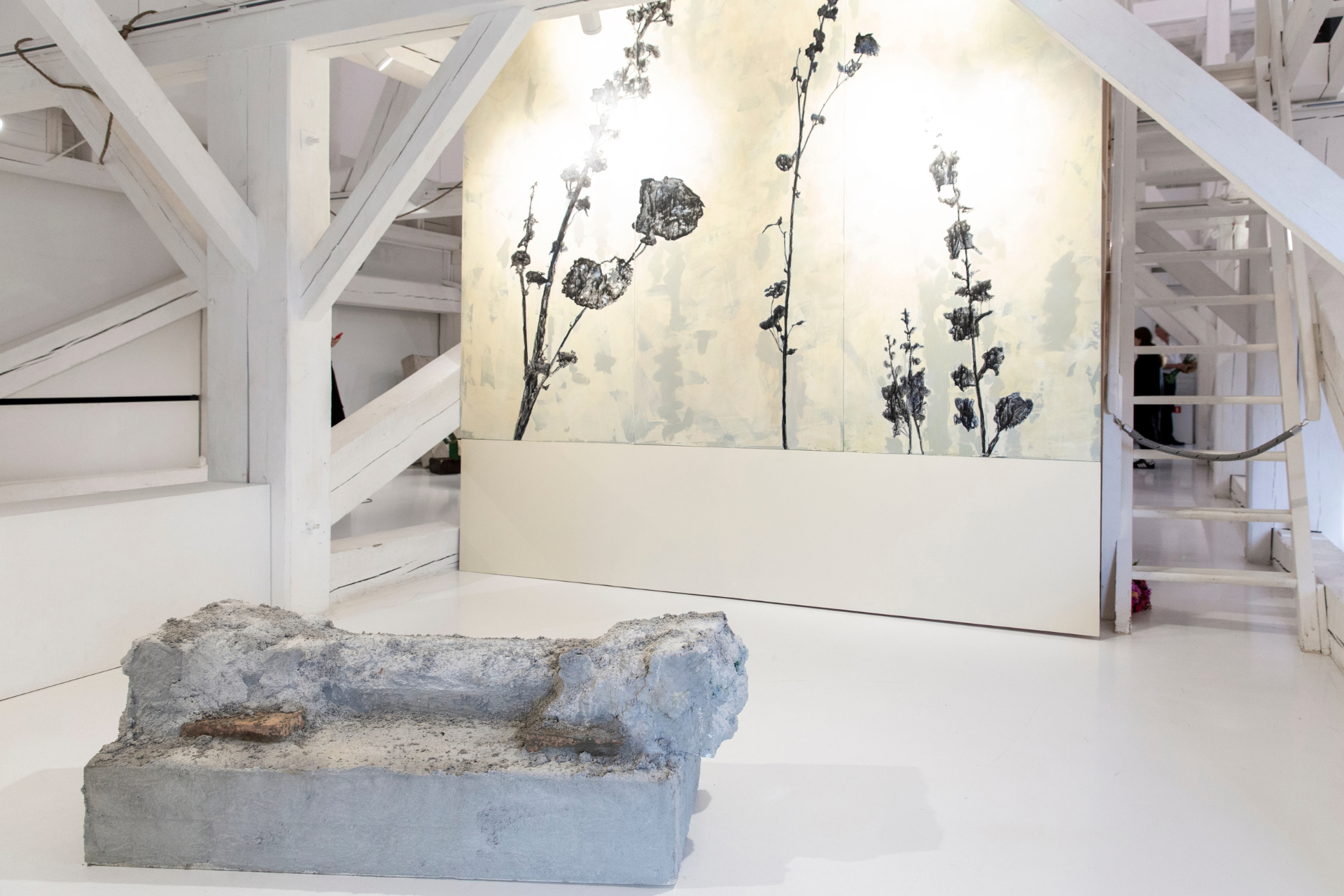 A contemporary art installation featuring a rough, gray concrete block at the forefront, with a white wooden staircase structure around it. Behind the staircase, there is a large panel of artwork depicting abstract black plant-like forms on a pale background. The setting appears to be a modern art gallery with white walls and floors.