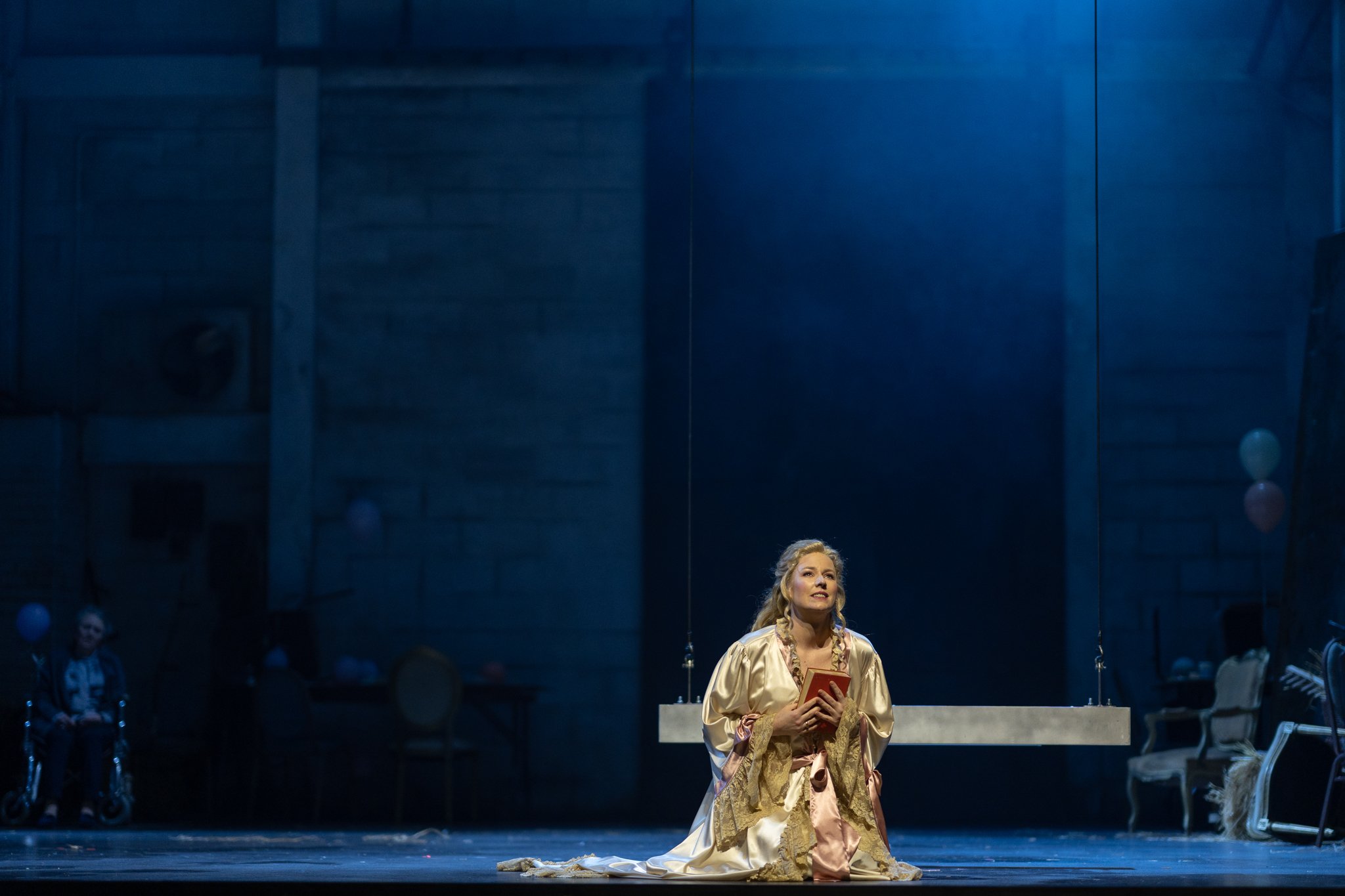 A woman in an elegant gown kneels on stage, holding a red book, with a joyful expression. The stage is dimly lit with blue hues, and several people are visible in the background, including a woman standing to the left with a person in a wheelchair and others standing or seated.