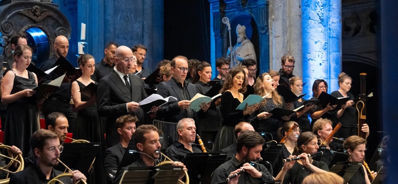 A choir and orchestra performing in a concert hall with blue lighting and classical sculptures in the background.