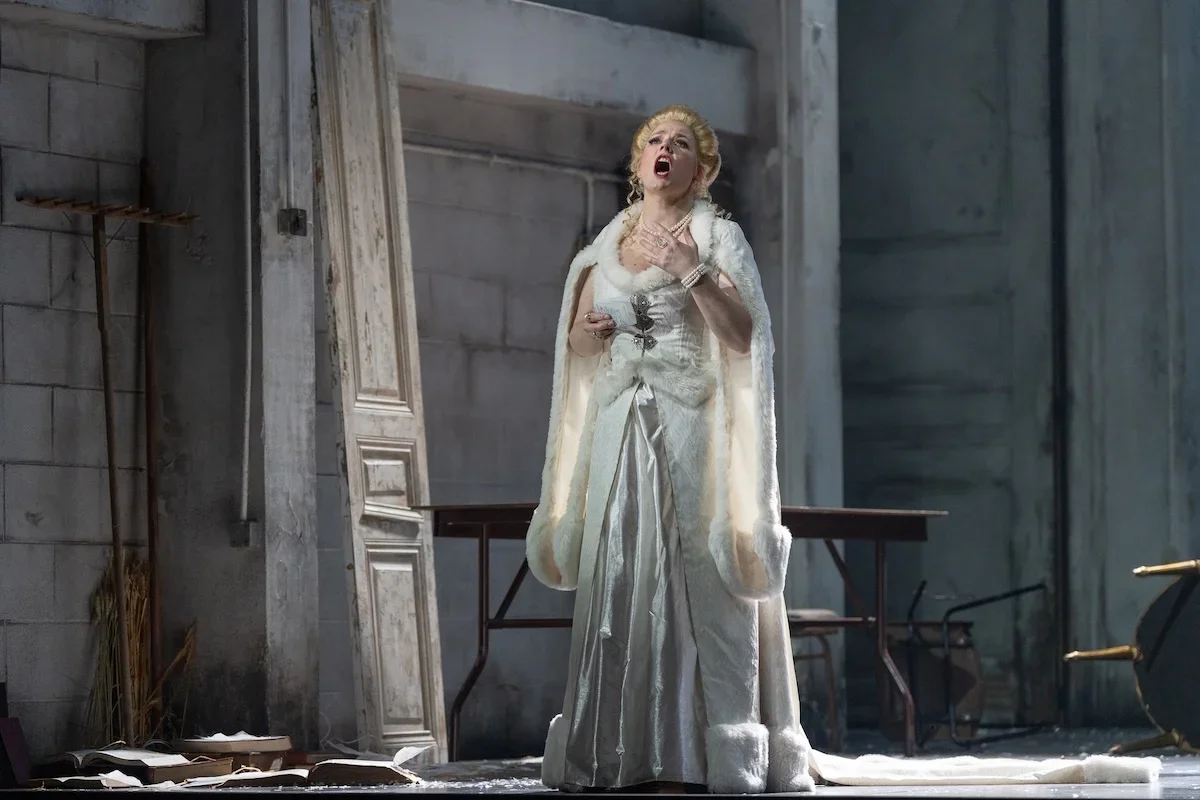 Woman dressed in an elegant white gown and fur coat, singing passionately on stage with a damaged, abandoned room setting.
