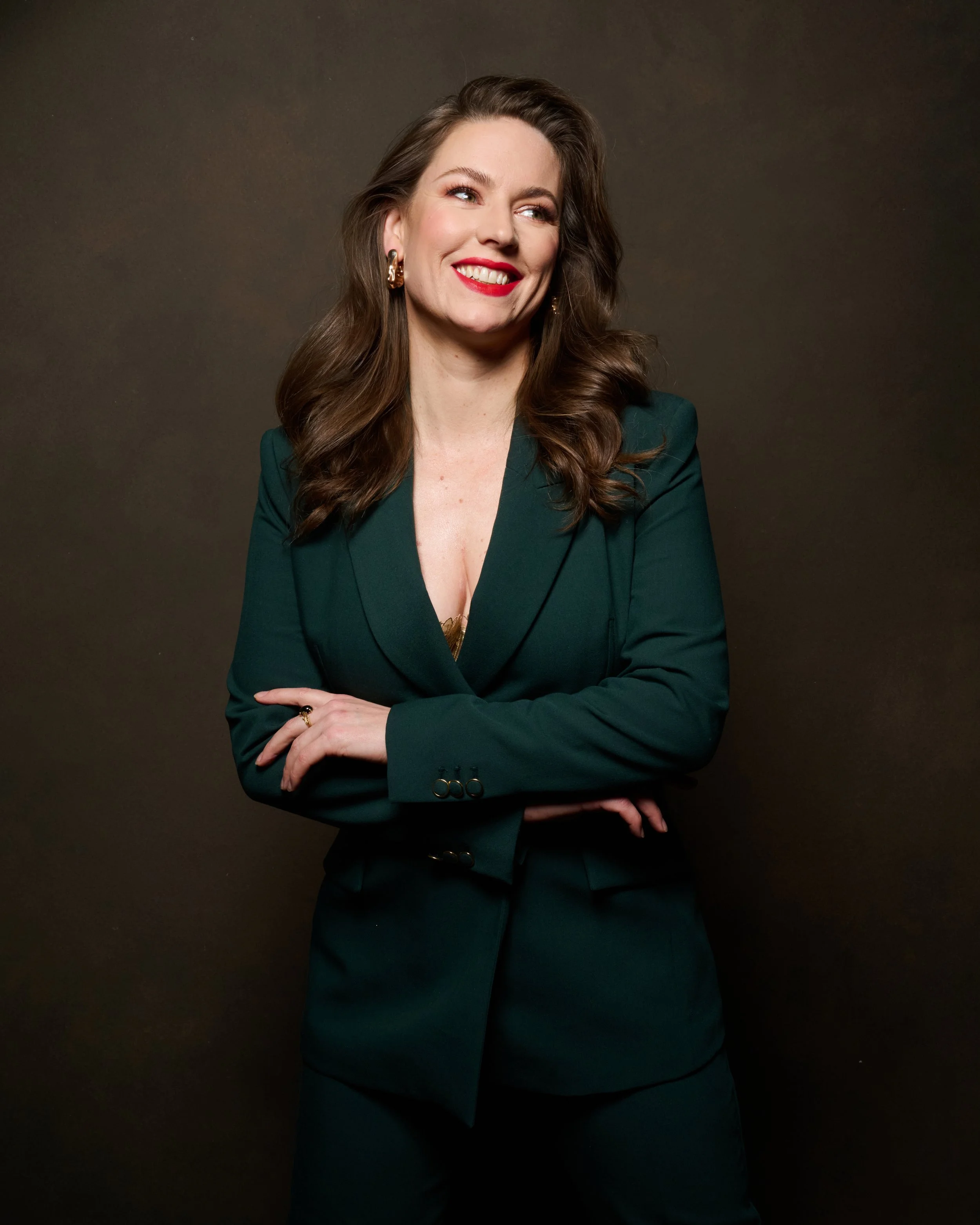 Smiling woman with brown hair, wearing a dark green suit and gold earrings, standing against a dark background.