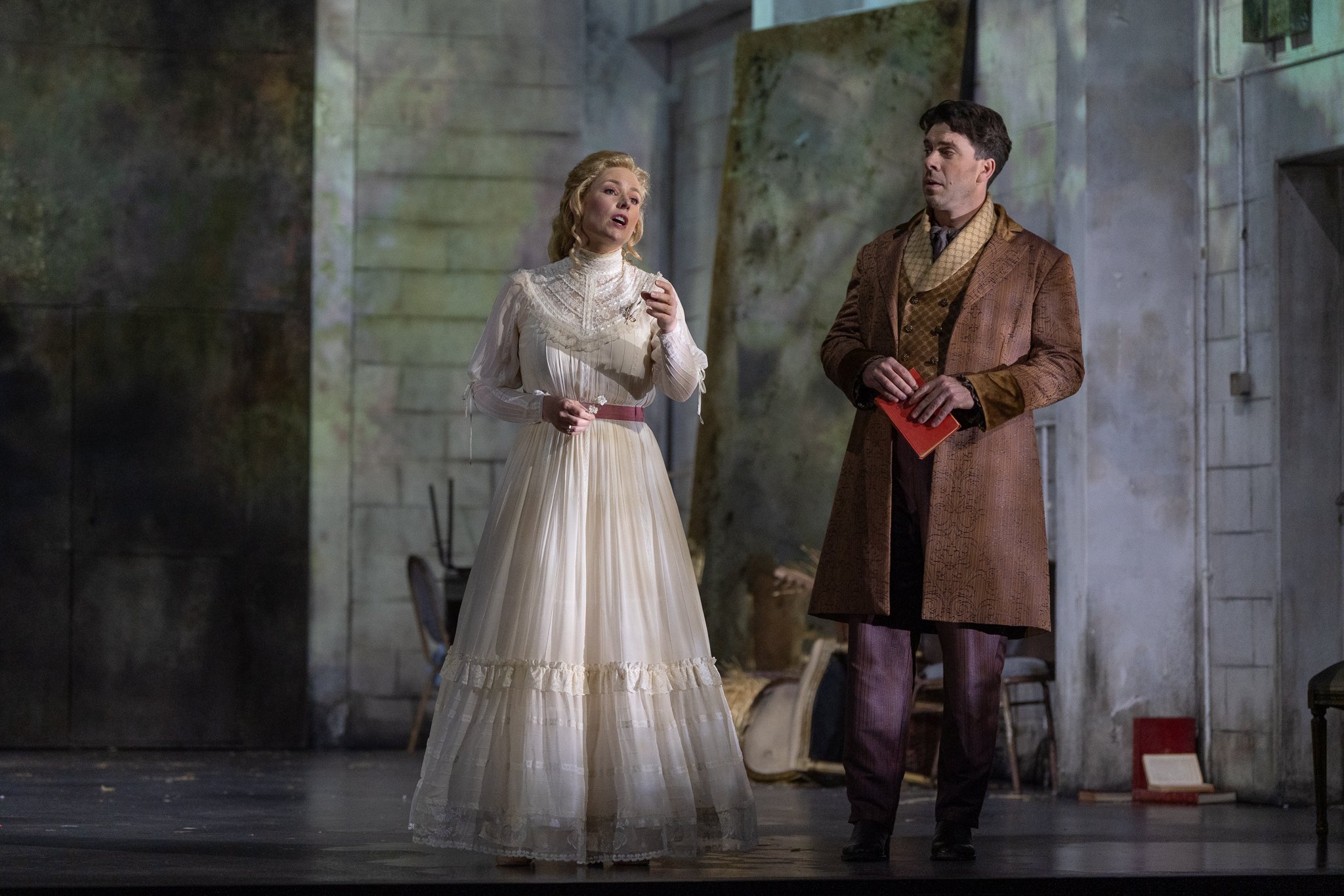 A scene from a theatrical play featuring a woman in a white vintage dress with a high neck and long sleeves, and a man in a brown patterned coat with a vest and cravat, standing on a stage with a stone wall backdrop, appearing to be in conversation.