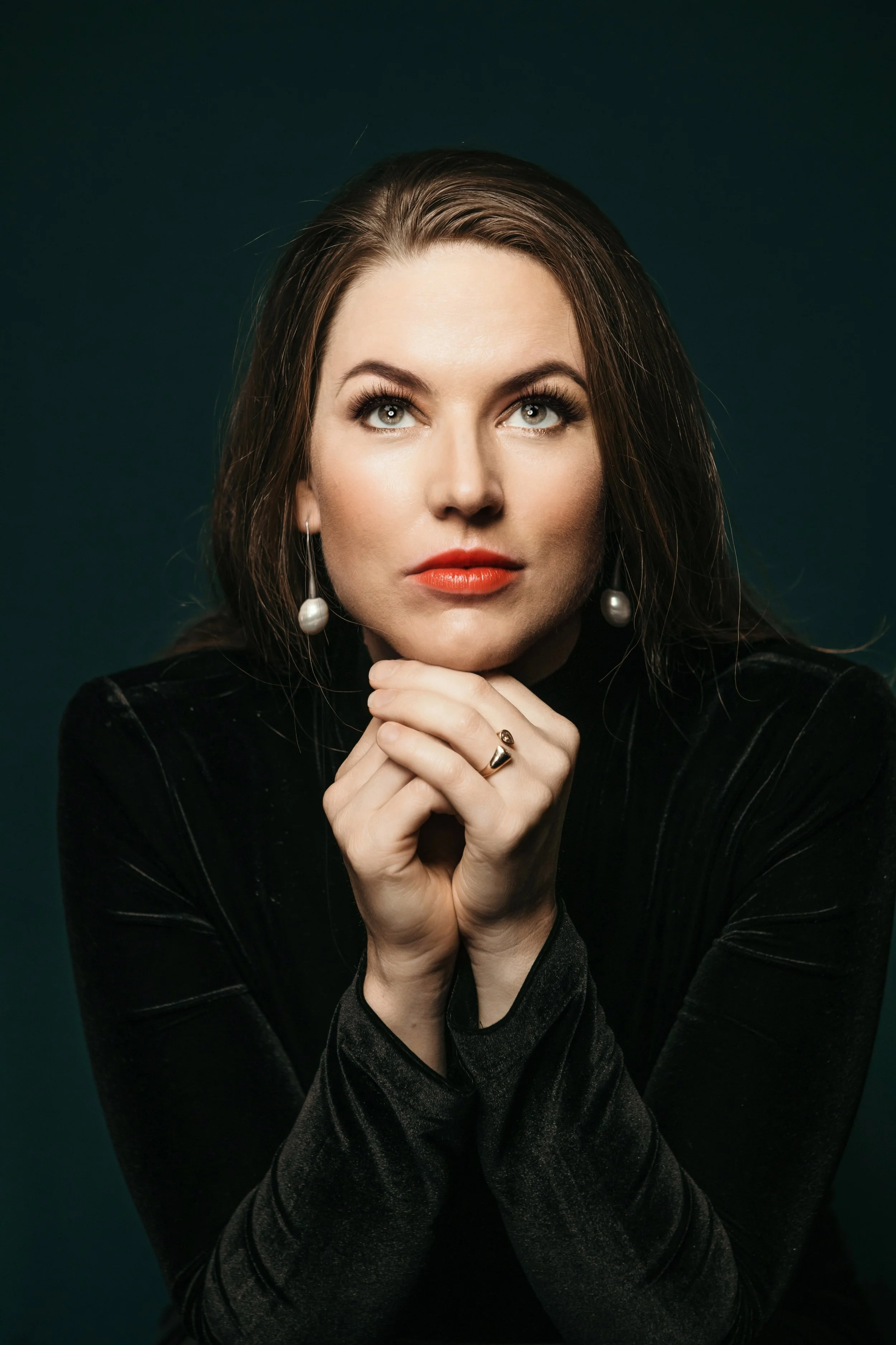 A woman with brown hair, fair skin, wearing pearl earrings and a black velvet top, poses with her hands under her chin against a dark green background.