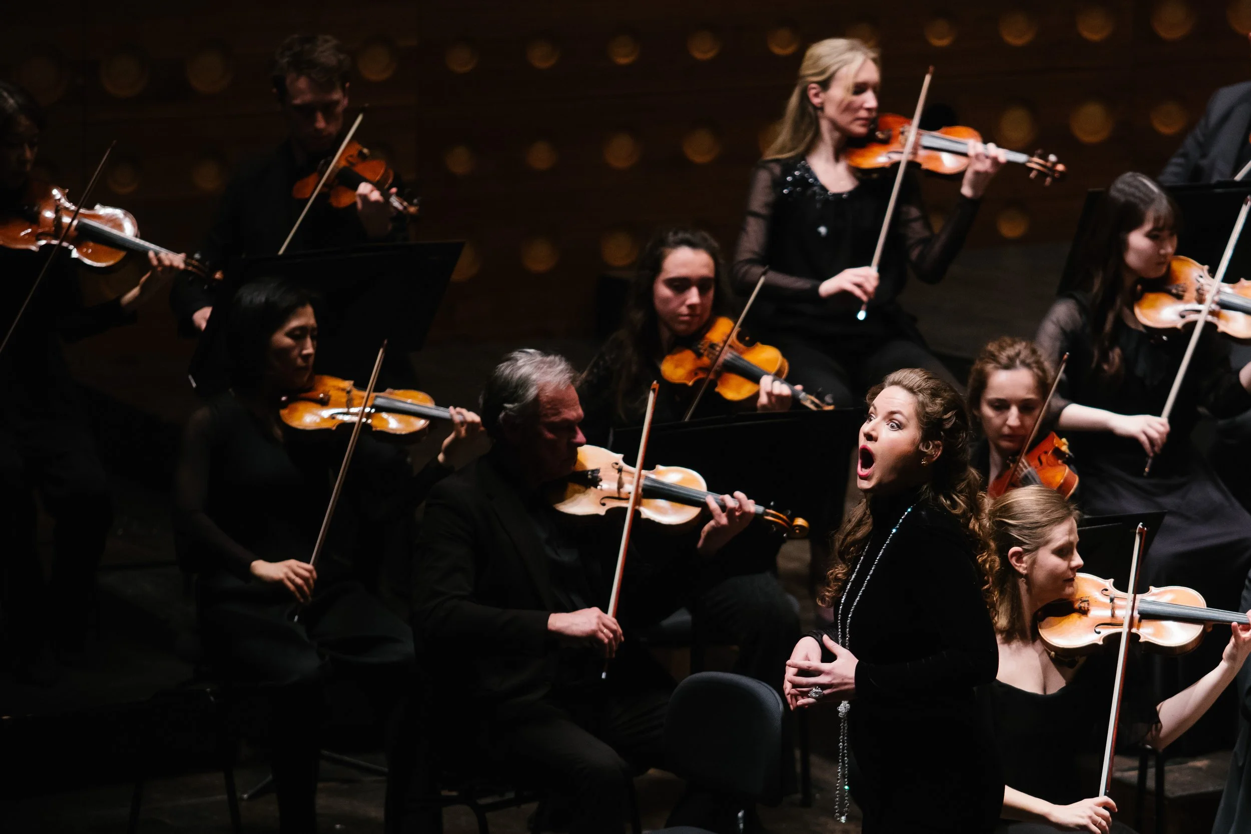Orchestra musicians playing violins during a performance, with a woman in the foreground singing or conducting, in a dimly lit concert hall.