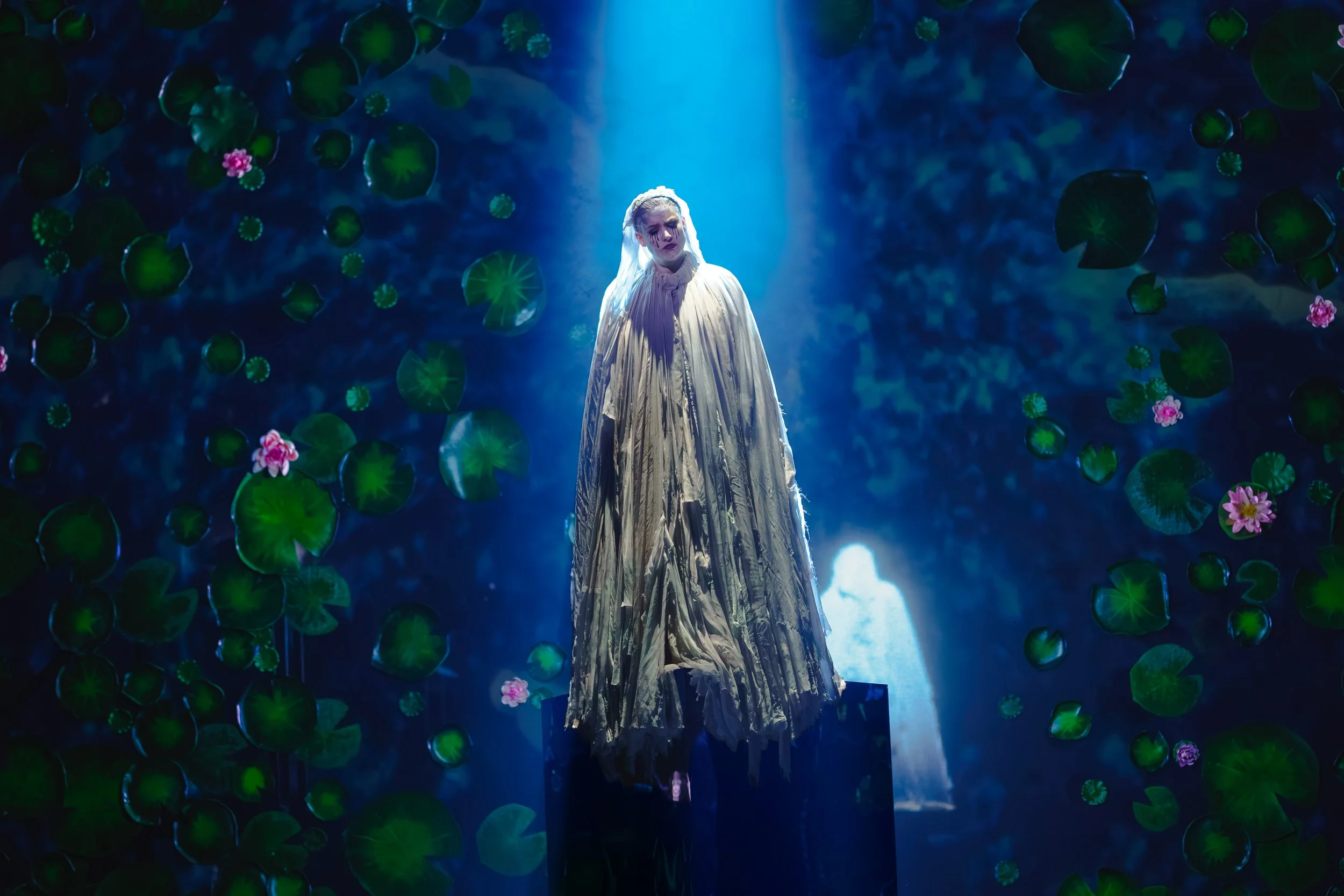 A woman dressed in a long, flowing, pale-colored gown stands on stage under a spotlight. The background is filled with floating green lily pads and pink water lilies, creating a surreal, dreamy atmosphere.