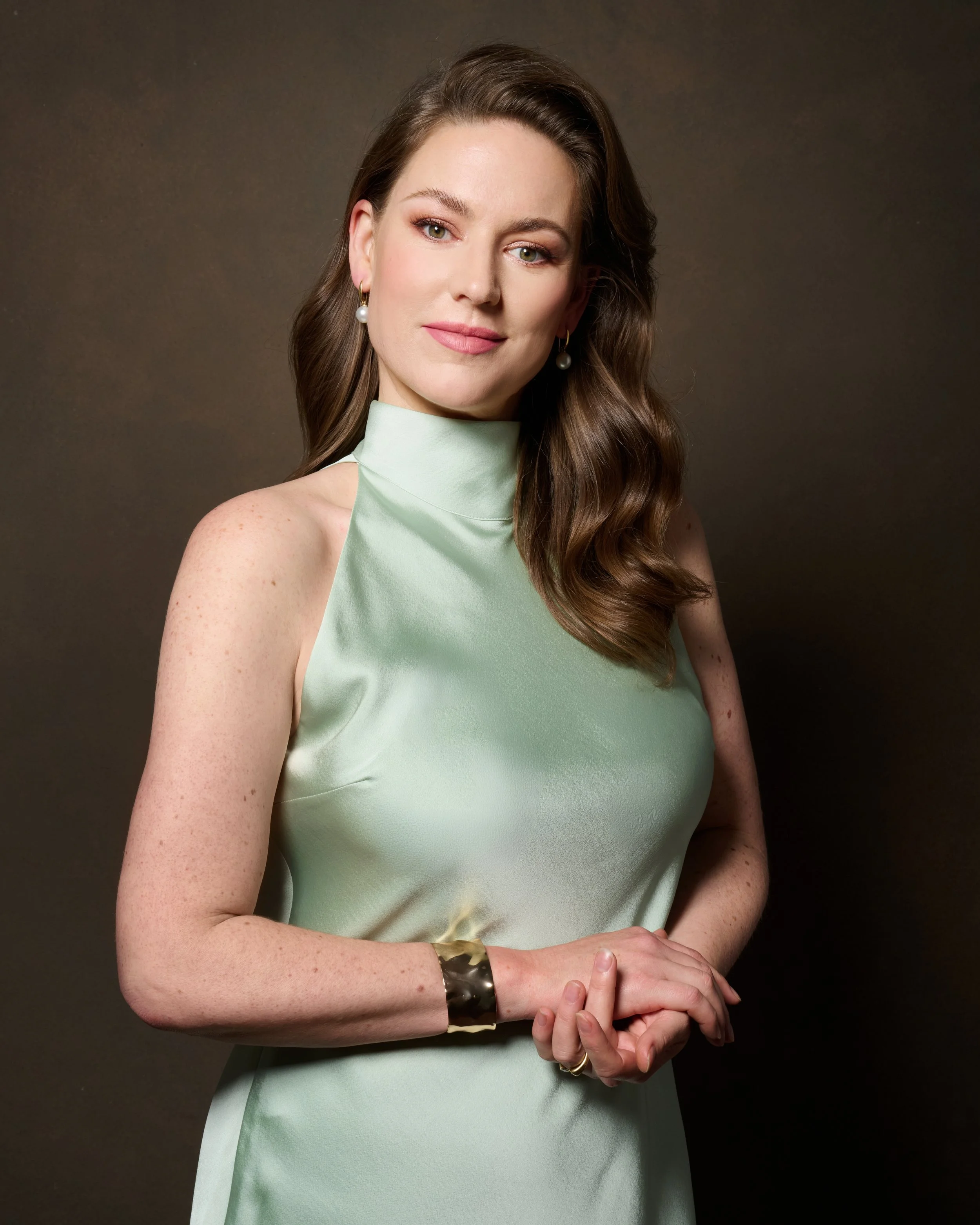 Portrait of a woman with long wavy brown hair, wearing pearl earrings, a light green sleeveless dress, silver bracelet, and a ring, standing against a dark background.