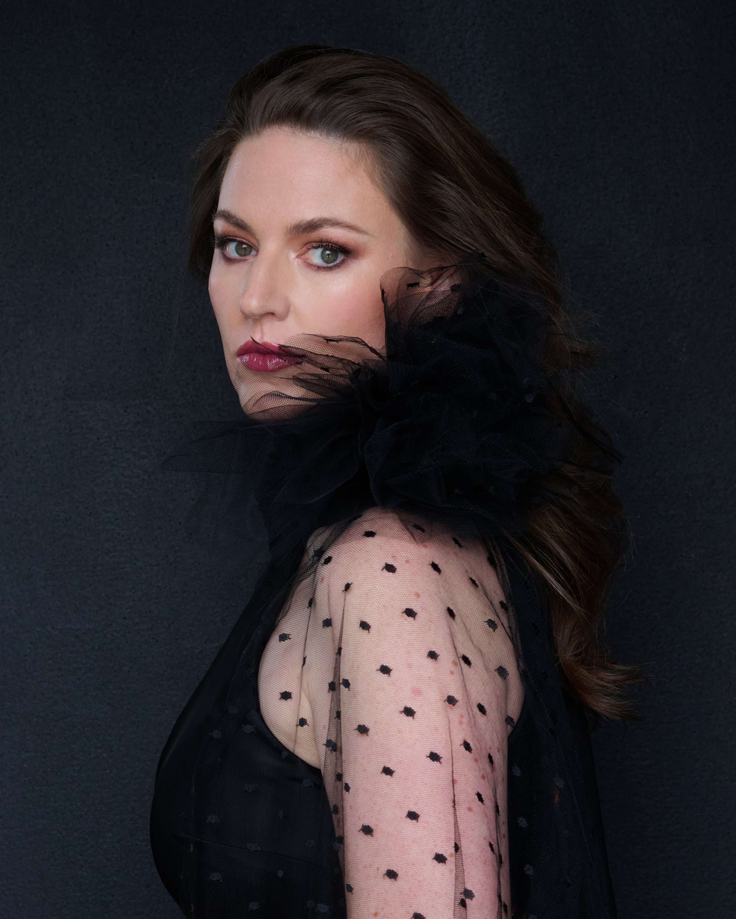 Eleanor Lyons with long brown hair and green eyes wearing a black polka dot sheer evening gown and black tulle fabric covering part of her face and shoulder, against a black background.
