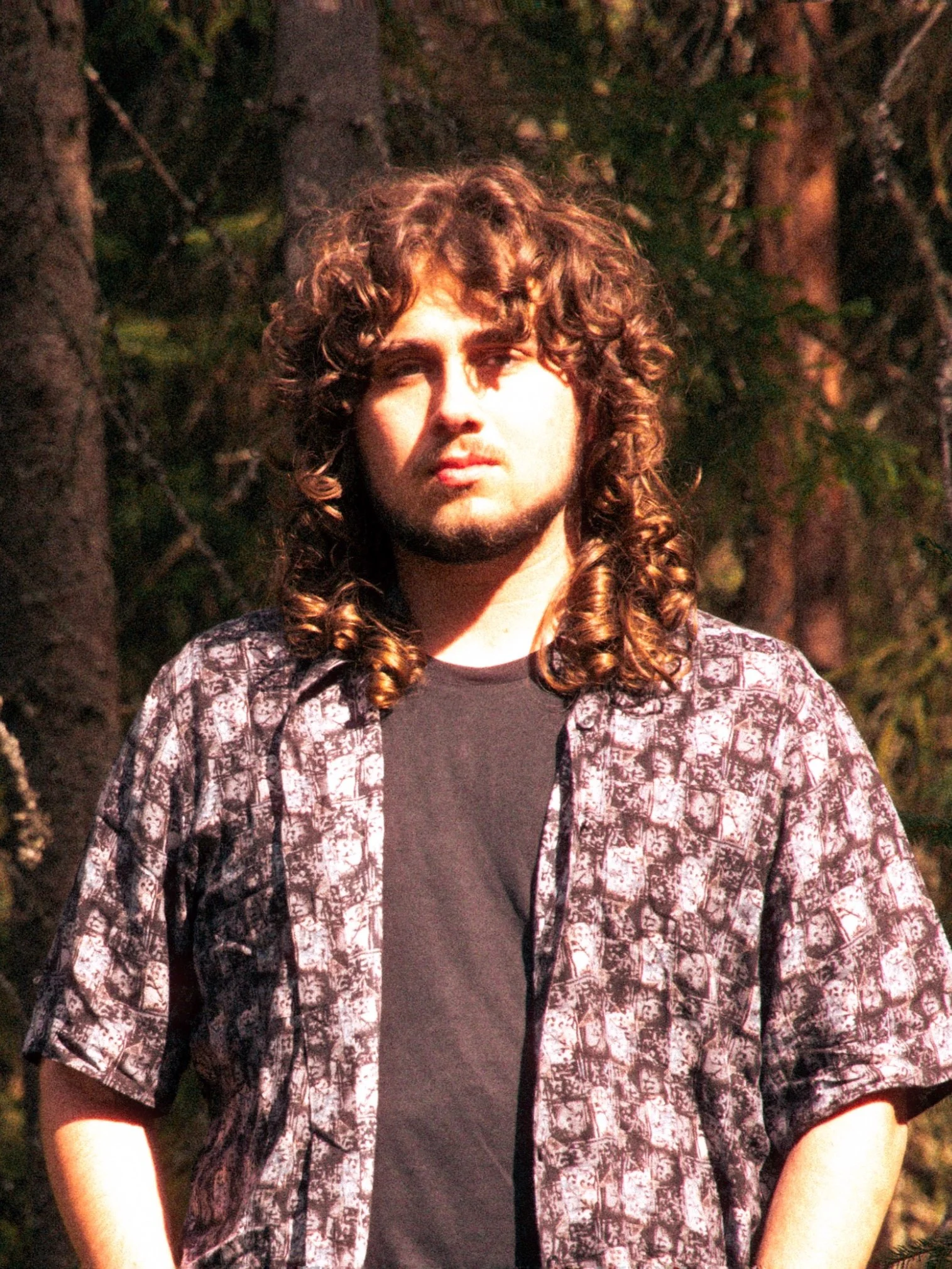 A man with curly brown hair and a beard, wearing a patterned shirt over a black t-shirt, standing outdoors among trees.