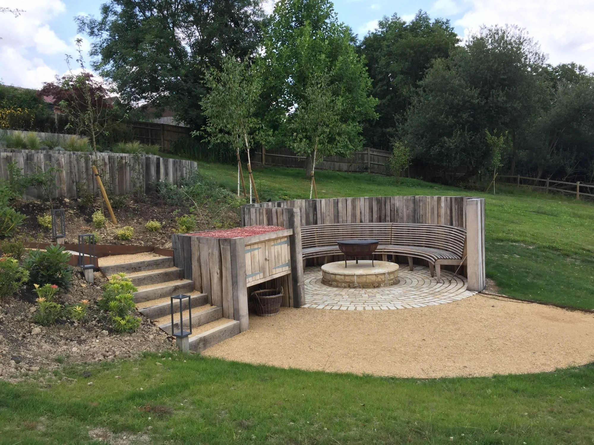 Finished curved oak bench around a fire bowl at Rotherhurst, with sleeper walls, circular paving and completed garden landscaping.