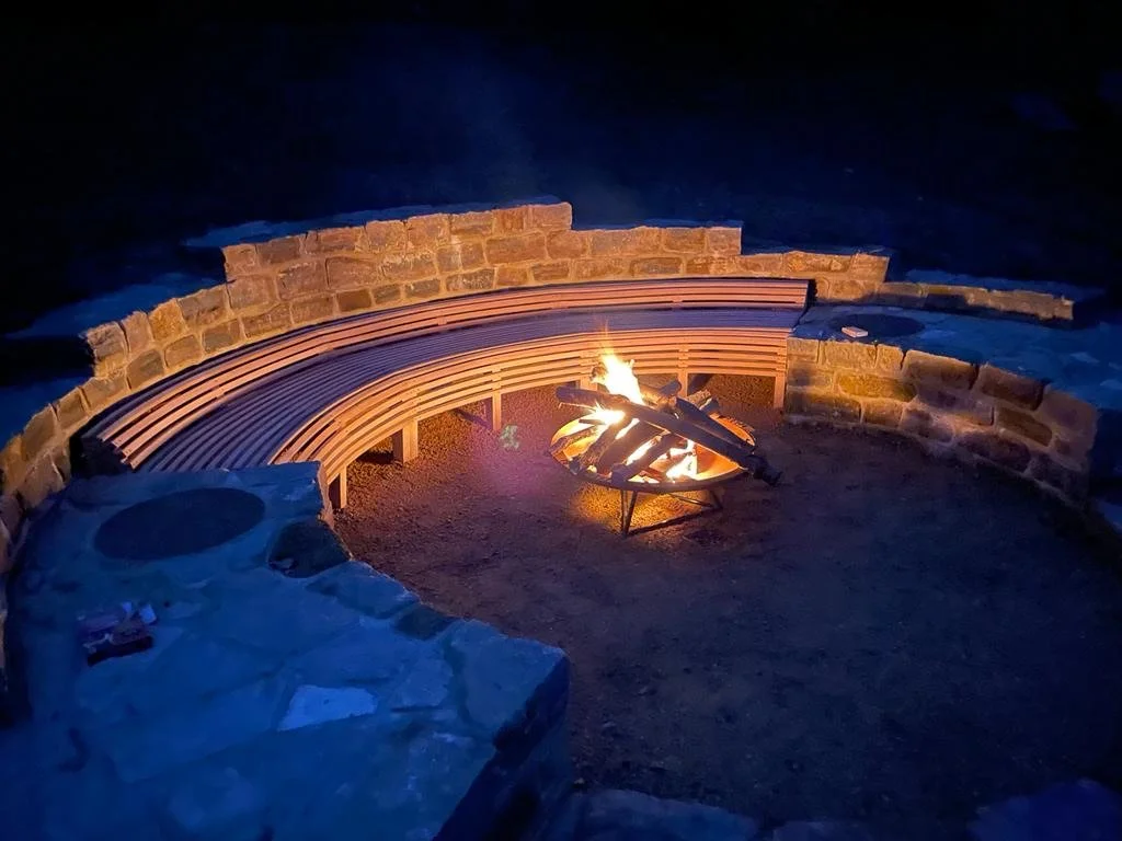 Holly Mount after dark, with curved oak seating, illuminated stone walls and a fire burning in the central bowl.
