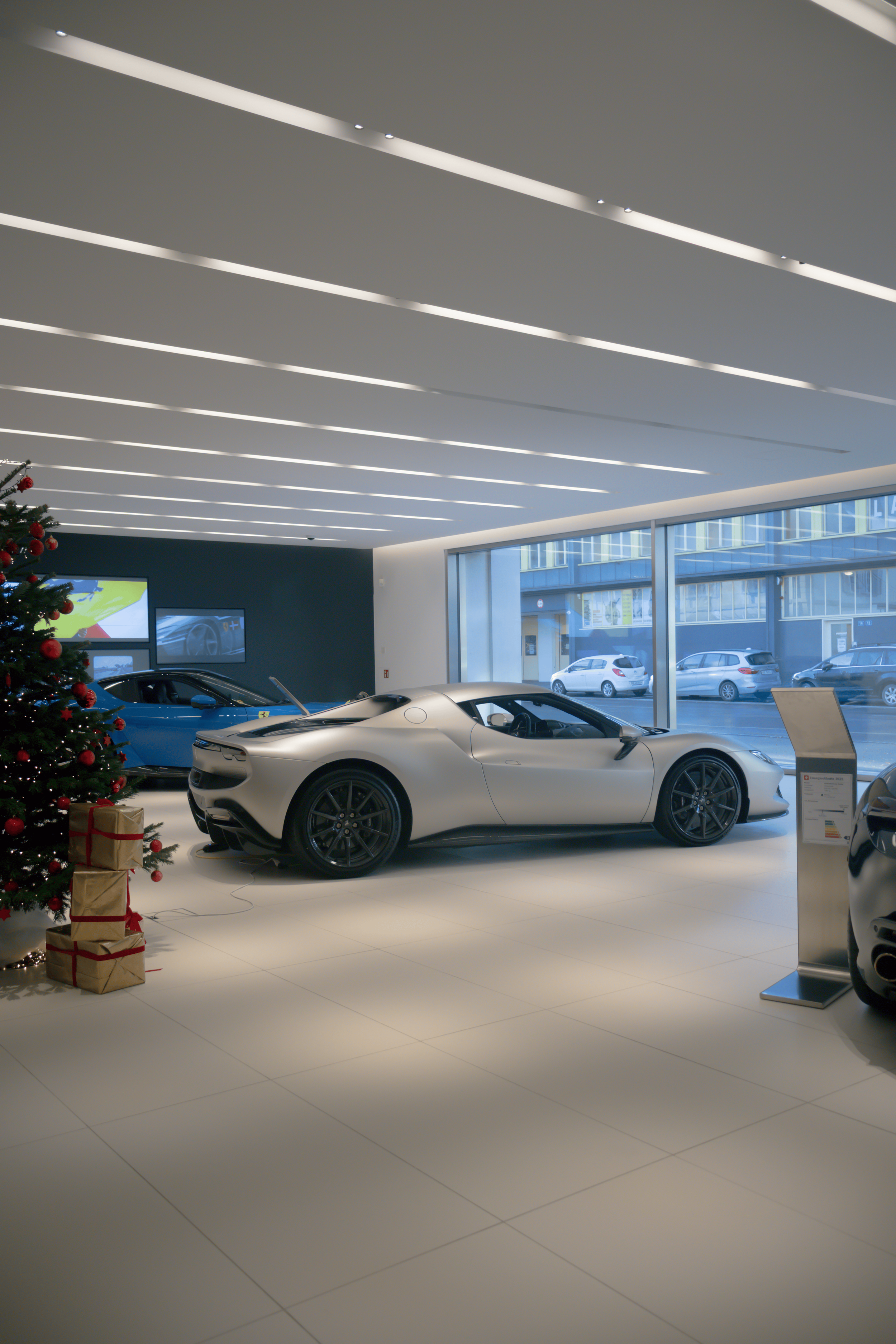 Inside a modern car dealership with white flooring, large glass windows, and several sports cars including a silver car in the foreground and a blue one in the background. There is a decorated Christmas tree with red ornaments and wrapped presents ne