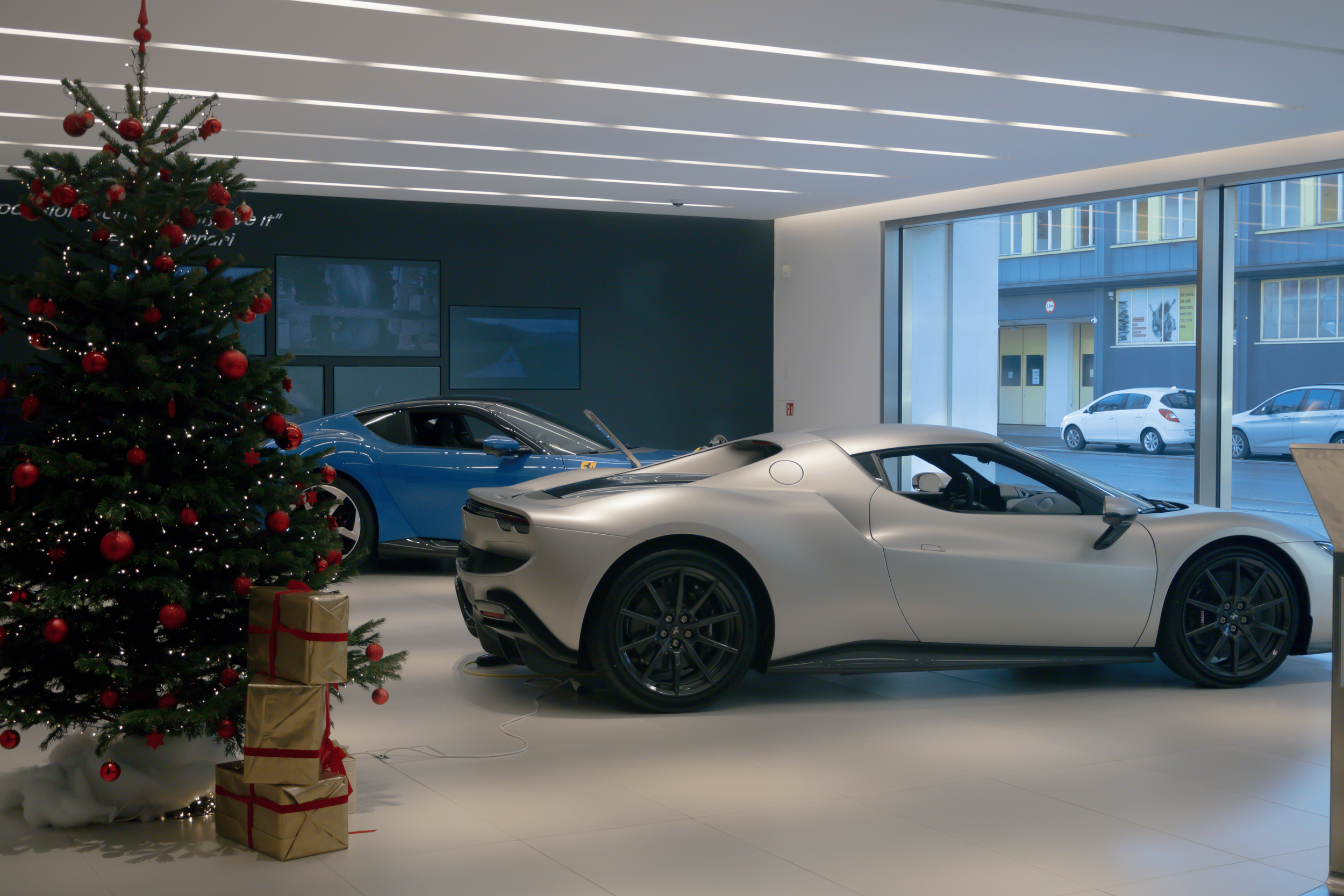 A Christmas tree decorated with red ornaments and lights, positioned next to luxury sports cars inside a modern car dealership with large glass windows showing parked cars outside.