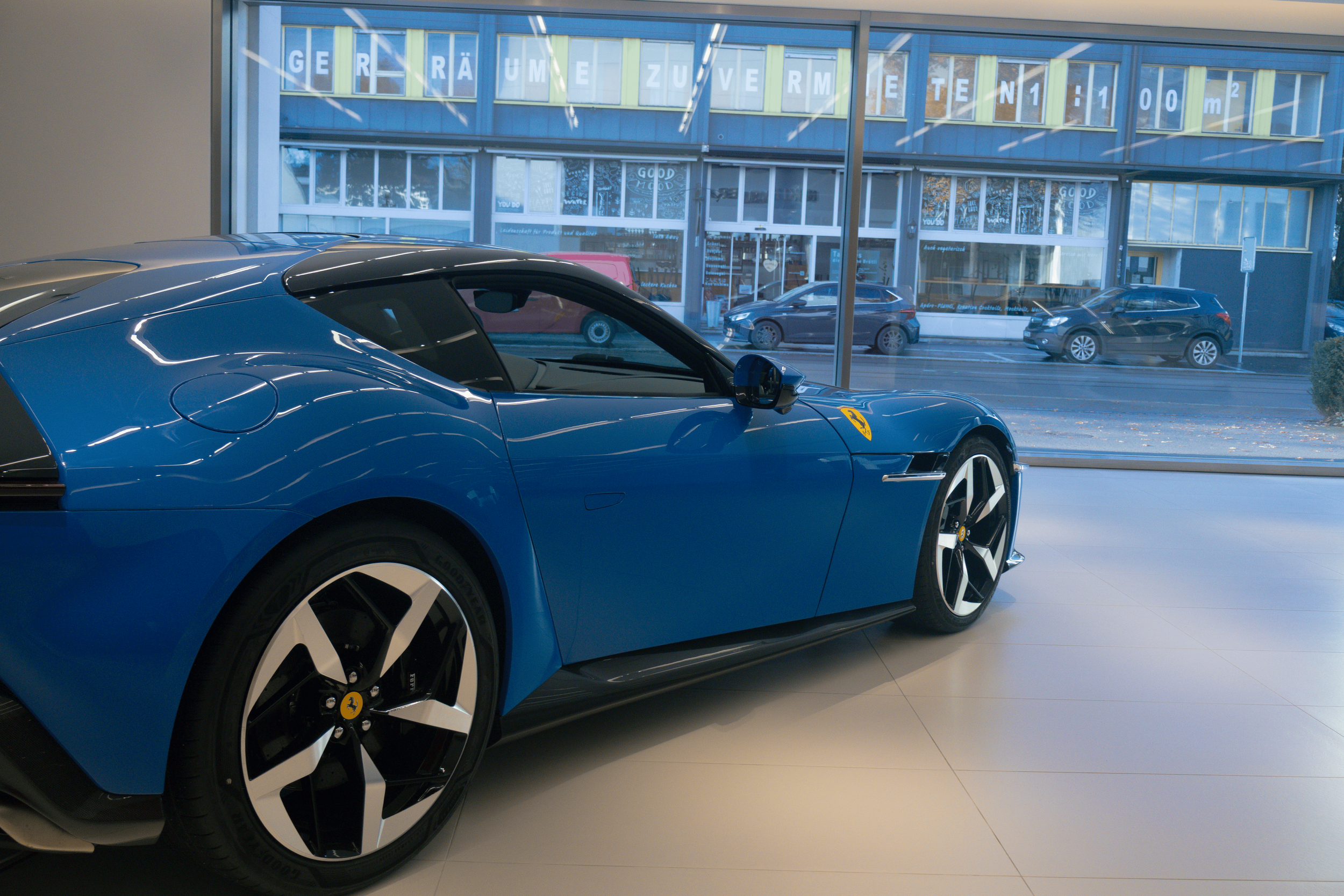 A blue Ferrari sports car inside a dealership showroom, with large glass windows in the background showing parked cars outside and a building with signs.
