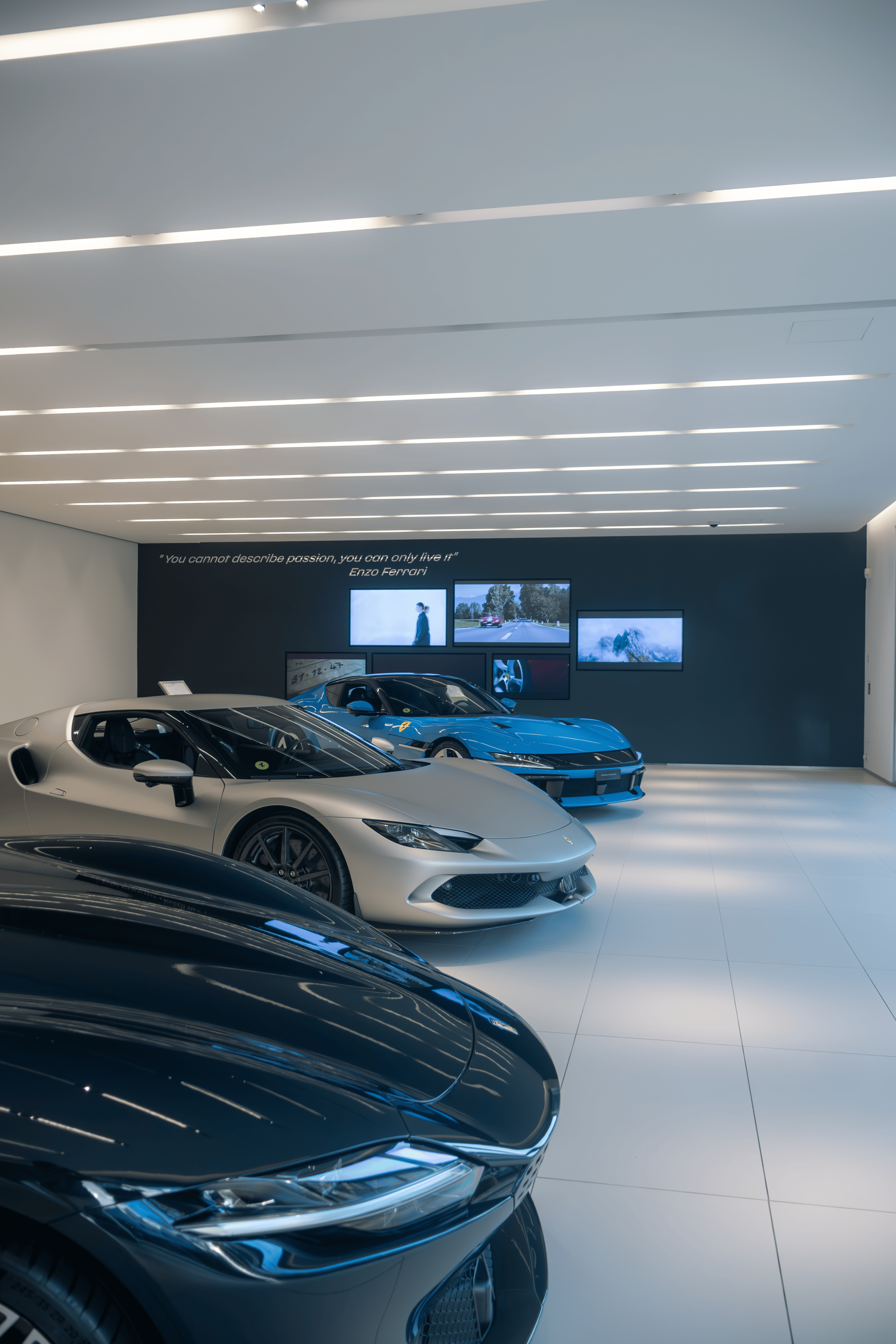 Luxury car showroom with a white sports car and two blue sports cars on display, featuring a quote by Enzo Ferrari on the black wall.