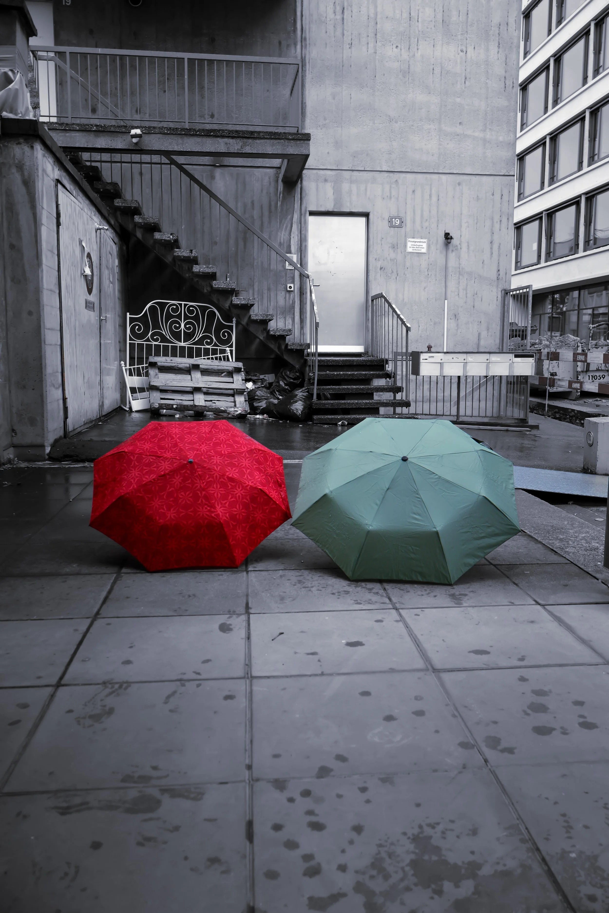 Three umbrellas in red, green, and blue are open on a wet sidewalk. The background shows a gray concrete building with a staircase, a door, and some construction material and fencing.