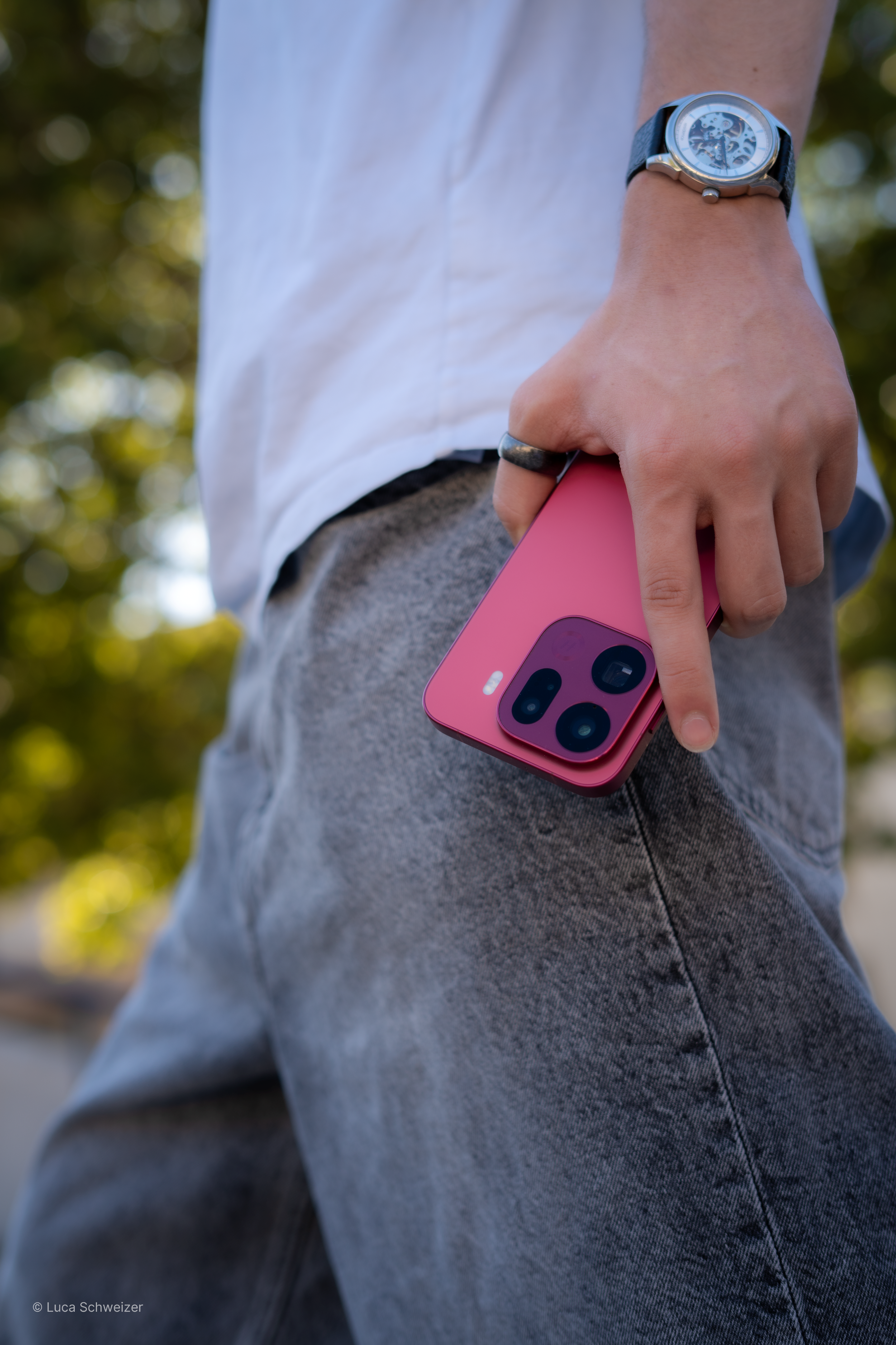 A person wearing a white shirt, gray pants, and a wristwatch with a round face and visible gears, balancing a pink smartphone between their thumb and middle finger. The background is blurred with green foliage.