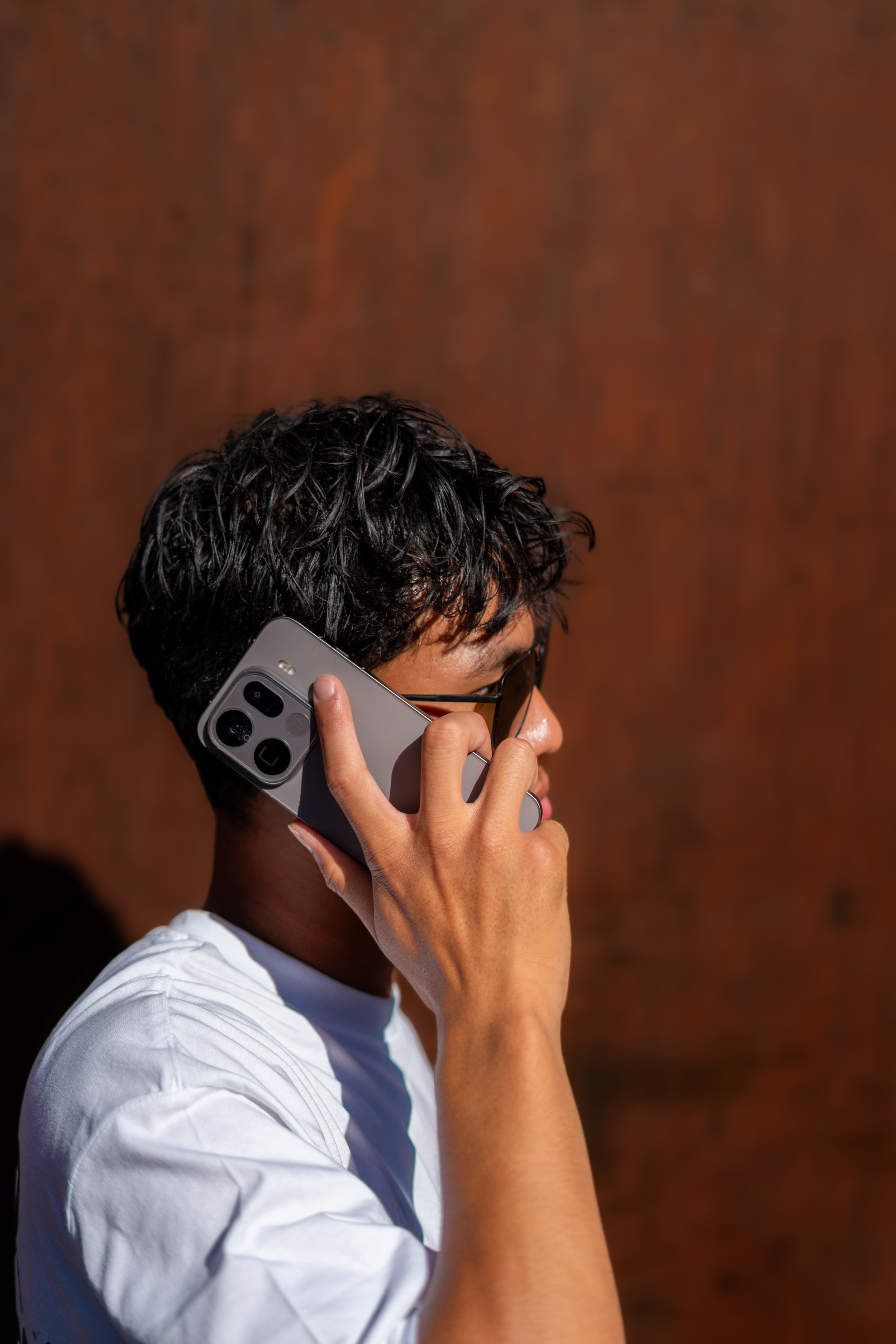 A person with dark, curly hair is holding a smartphone to their ear, wearing glasses and a white shirt. The background is a plain brown surface.