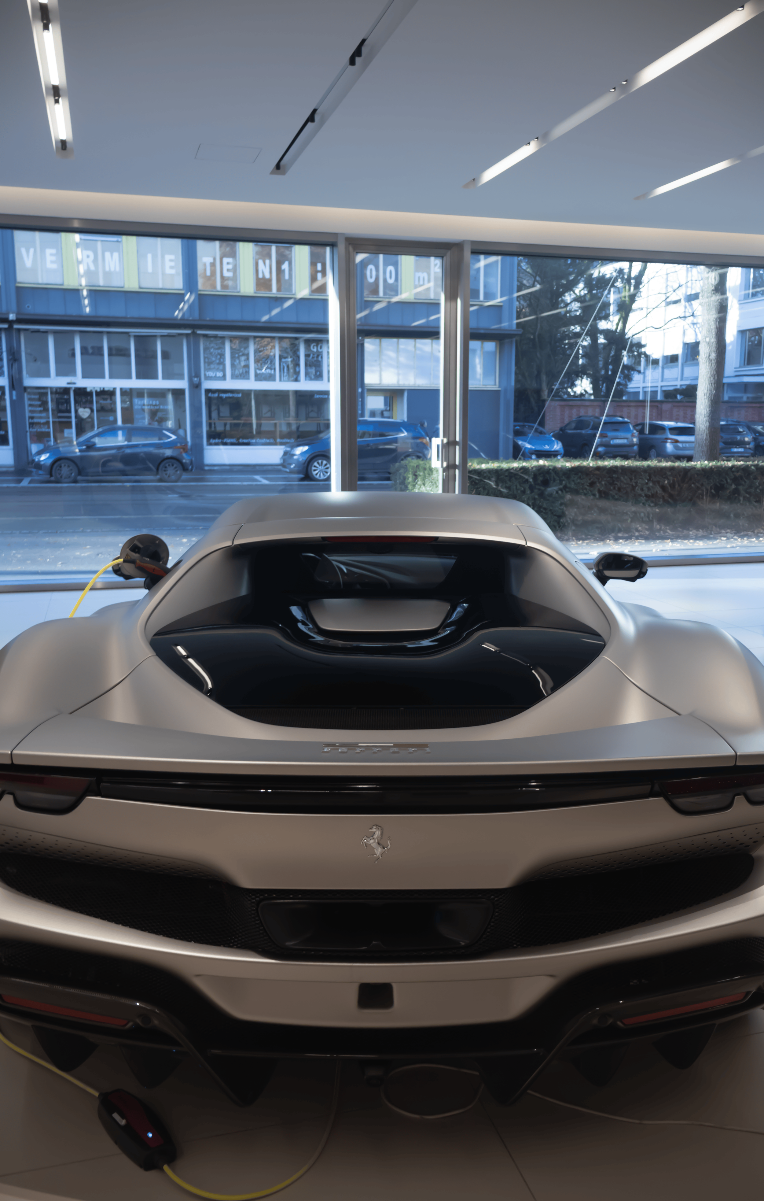 The image shows the rear view of a white sports car, possibly a Ferrari, inside a showroom with large windows. The car is plugged in for charging, with a charging cable connected to the left rear side.