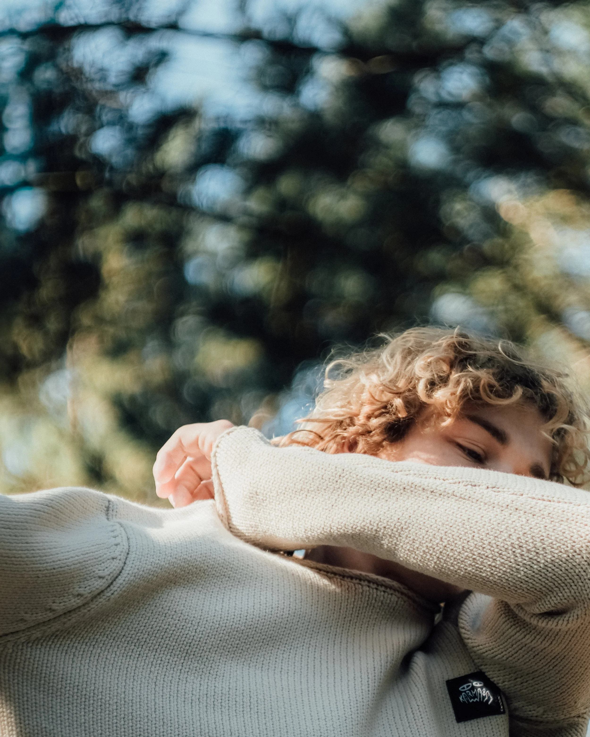 Enfant avec des cheveux bouclés, portant un pull beige, faisant un bras d'honneur, fond d'arbres flous.