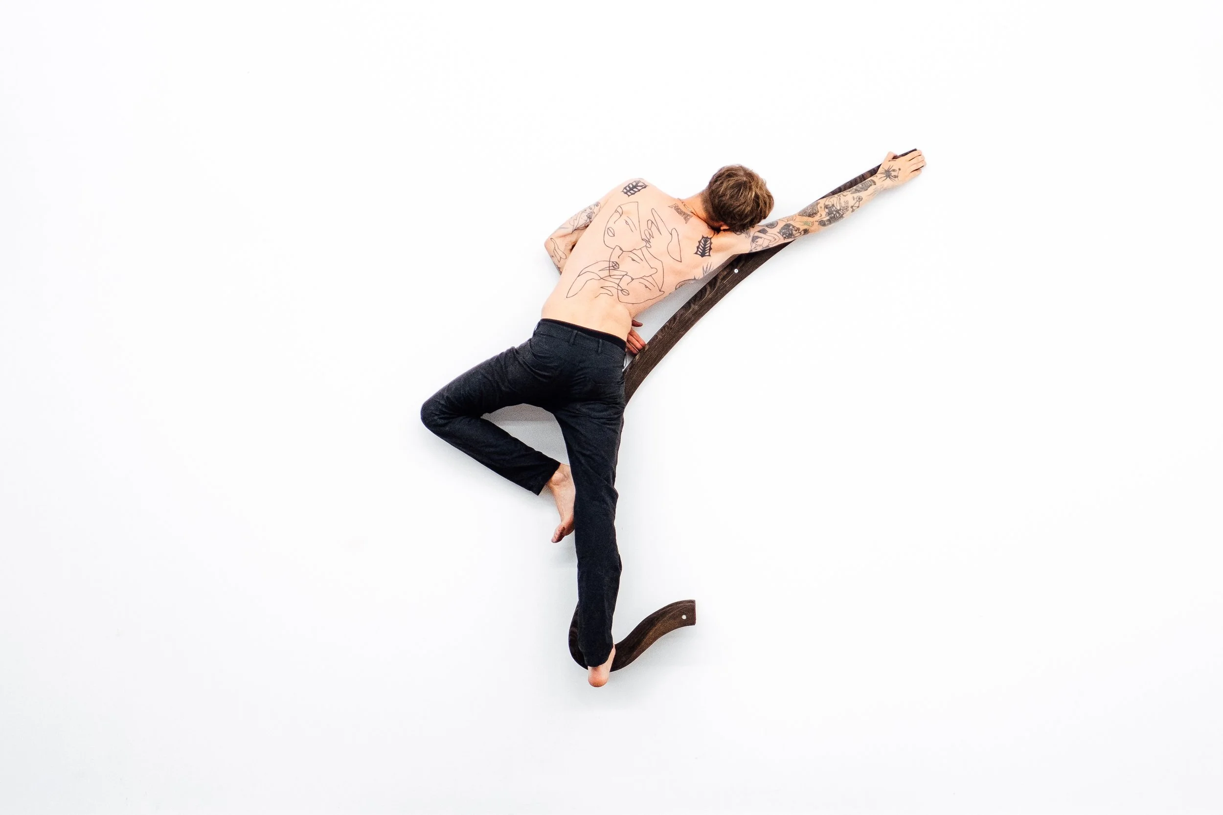 Jeune homme nu avec tatouages allongé sur un mur blanc, utilisant une scie en bois comme support