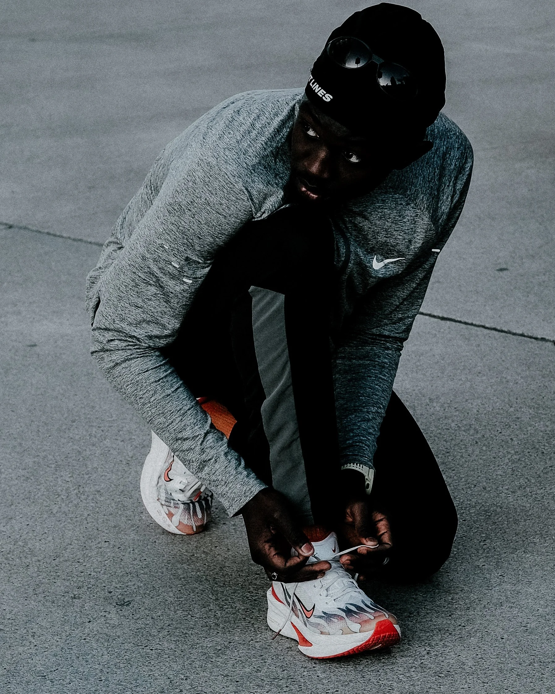 Une personne en tenue de sport, portant un bonnet noir avec des lunettes de soleil, est assise sur le sol en train de lacer ses chaussures de course. La personne porte un sweatshirt gris, un pantalon noir et des baskets blanches avec des motifs rouge