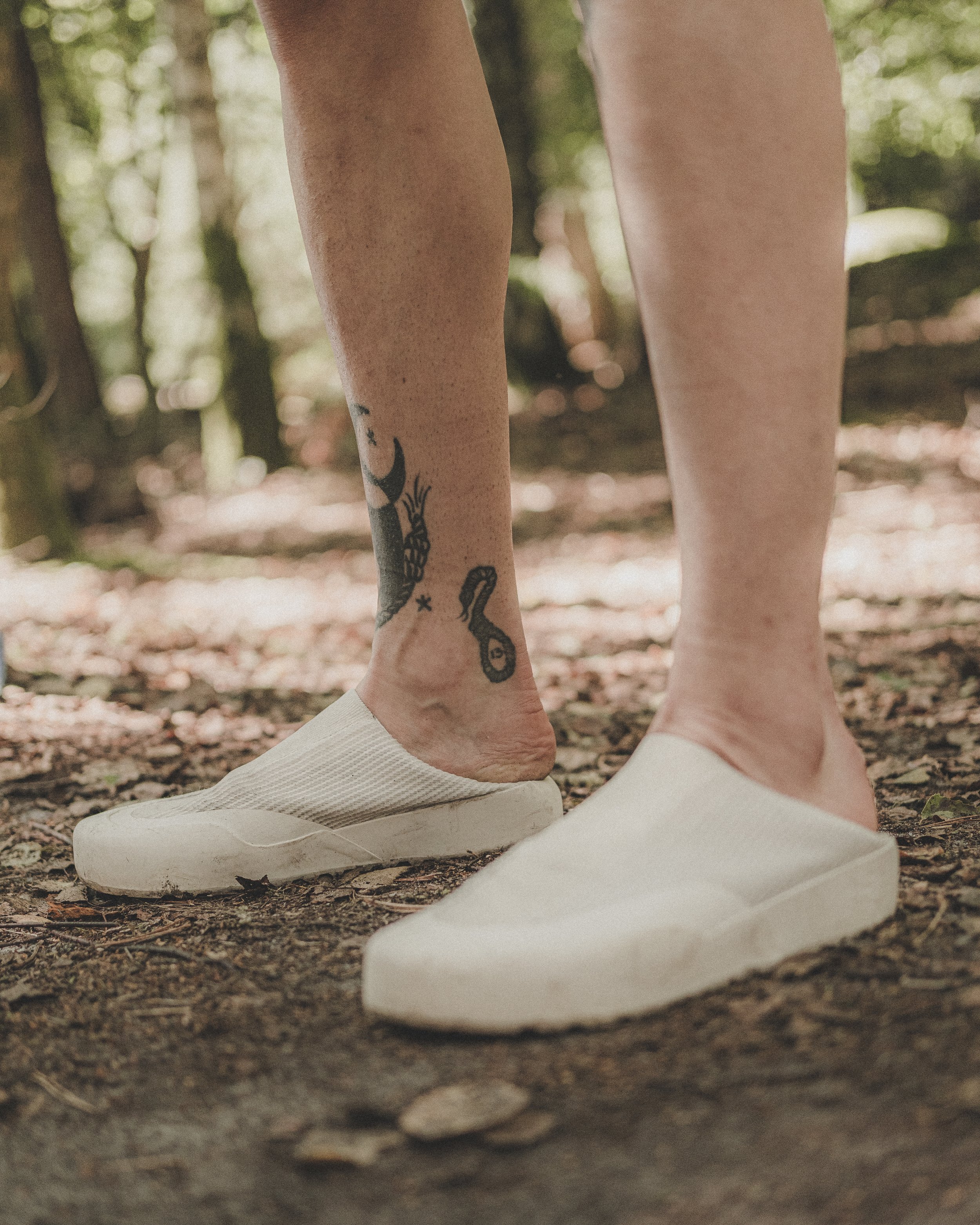 Les jambes d'une personne portant des chaussures blanches et un tatouage visible sur la jambe gauche, debout sur une surface de terre dans une forêt, entourée d'arbres et de feuilles.