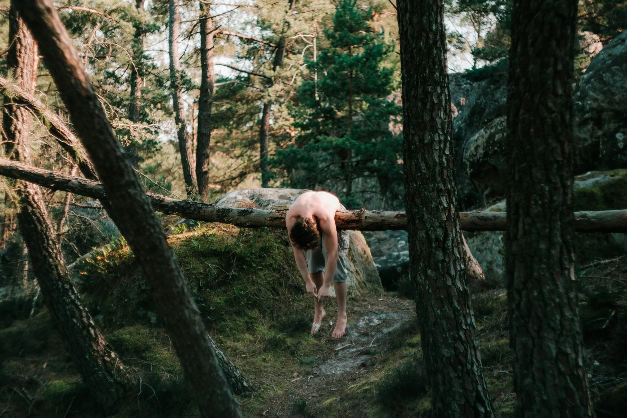 Une personne nue penchée en avant dans une forêt de pin, avec des arbres et des rochers autour, dans un environnement naturel.