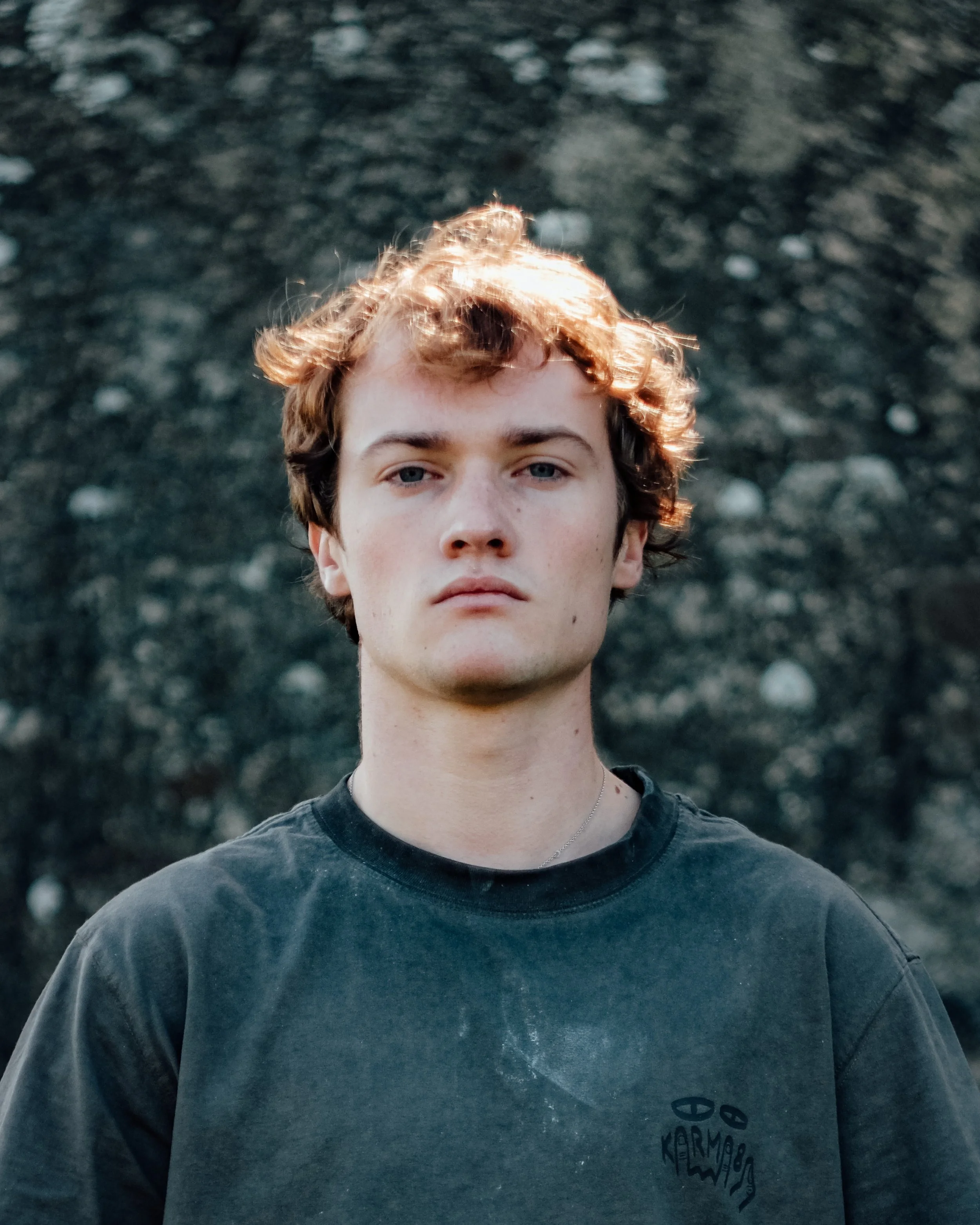 Un jeune homme avec des cheveux bouclés et blonds, portant un t-Shirt gris, se tient devant un fond rocheux, photo prise en plein air.