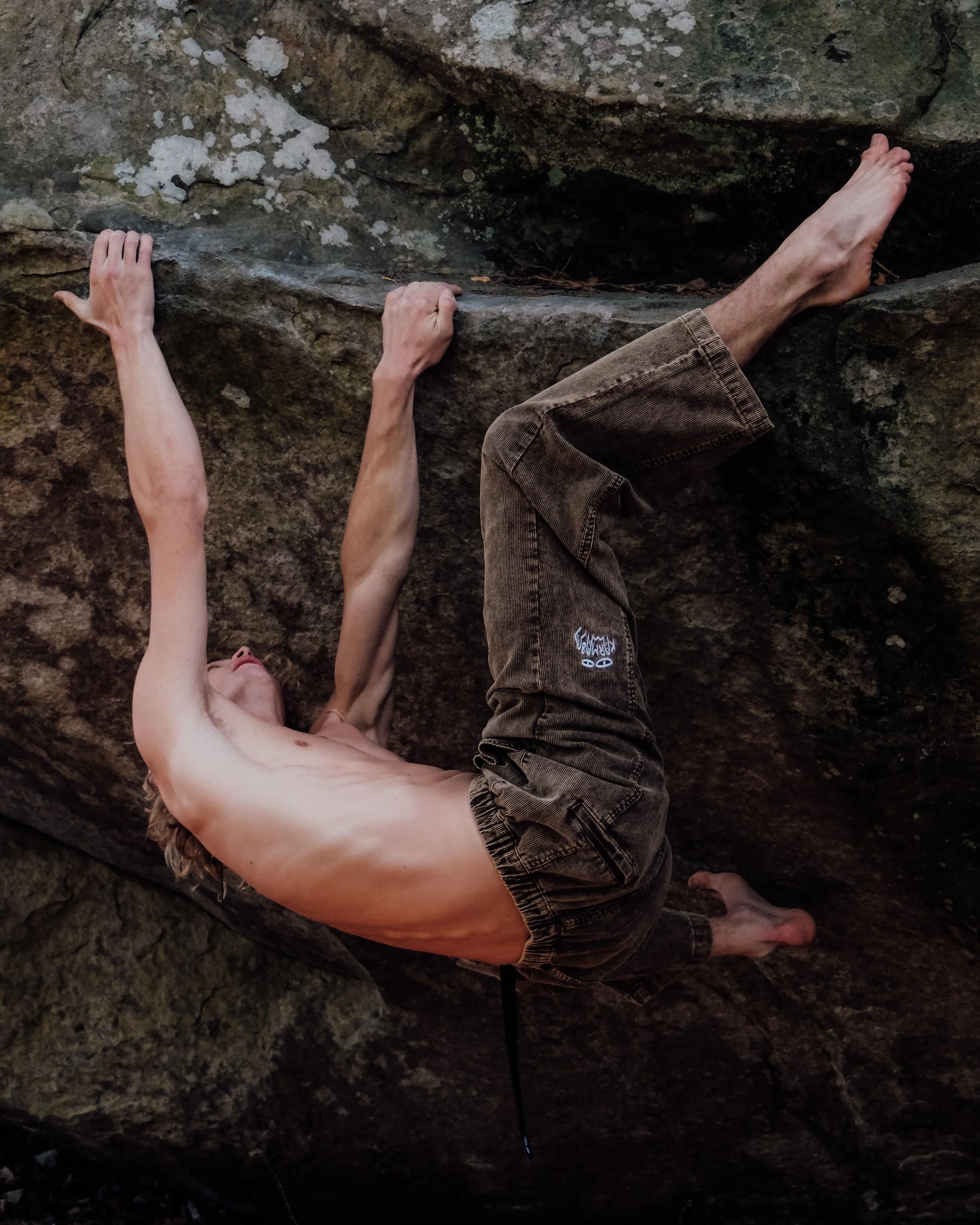 Un homme nu faisant de l'escalade en montagne rocheuse, vue de côté, tenant une prise avec une main et l'autre tendue vers une autre prise, portant un pantalon en velours côtelé. Il est attaché à une corde d'escalade noire.