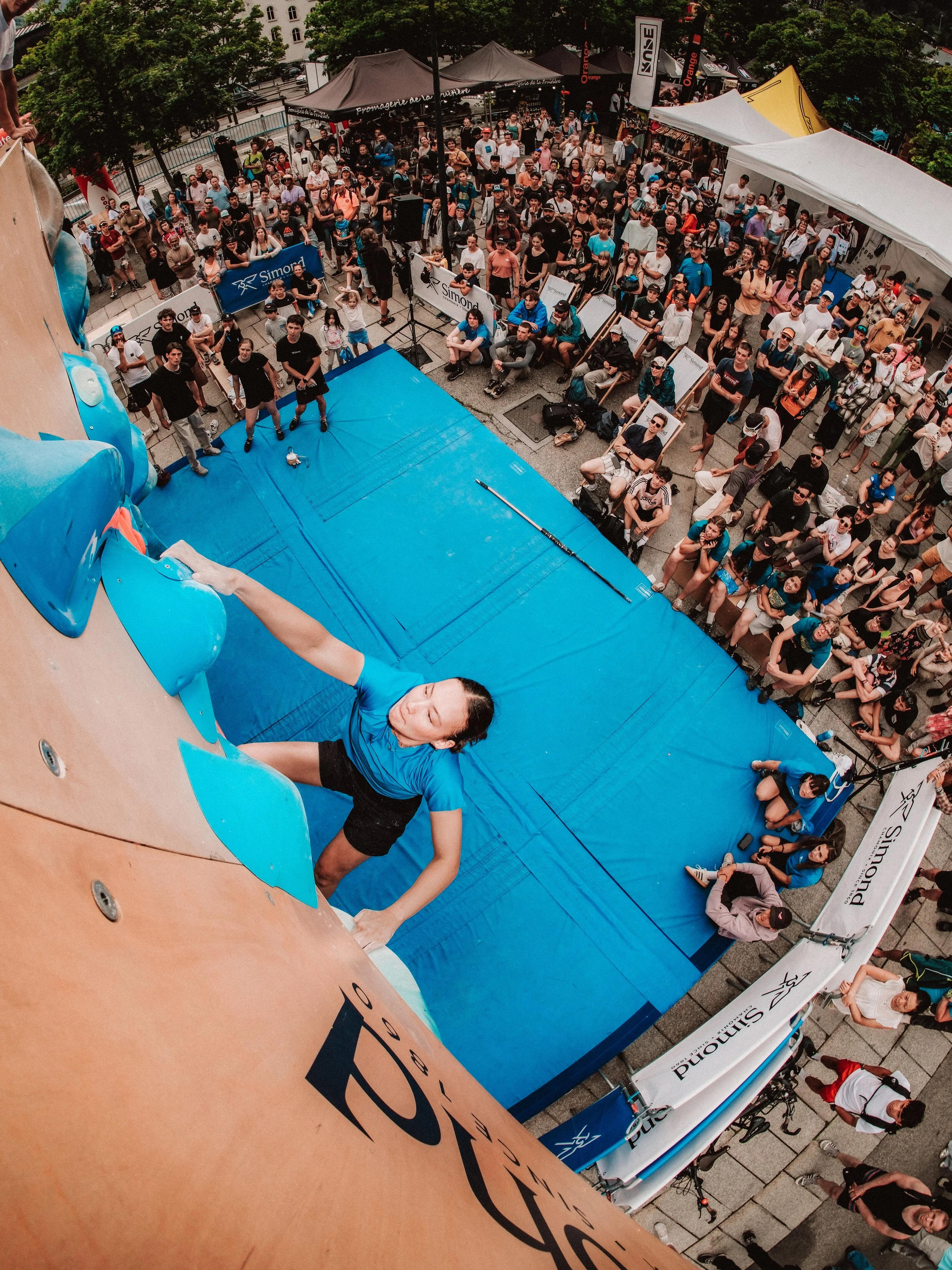 Une femme grimpe une paroi d'escalade en extérieur, vue d'en haut avec une foule de spectateurs regardant, en partie couverte par une bâche bleue et des tentes.