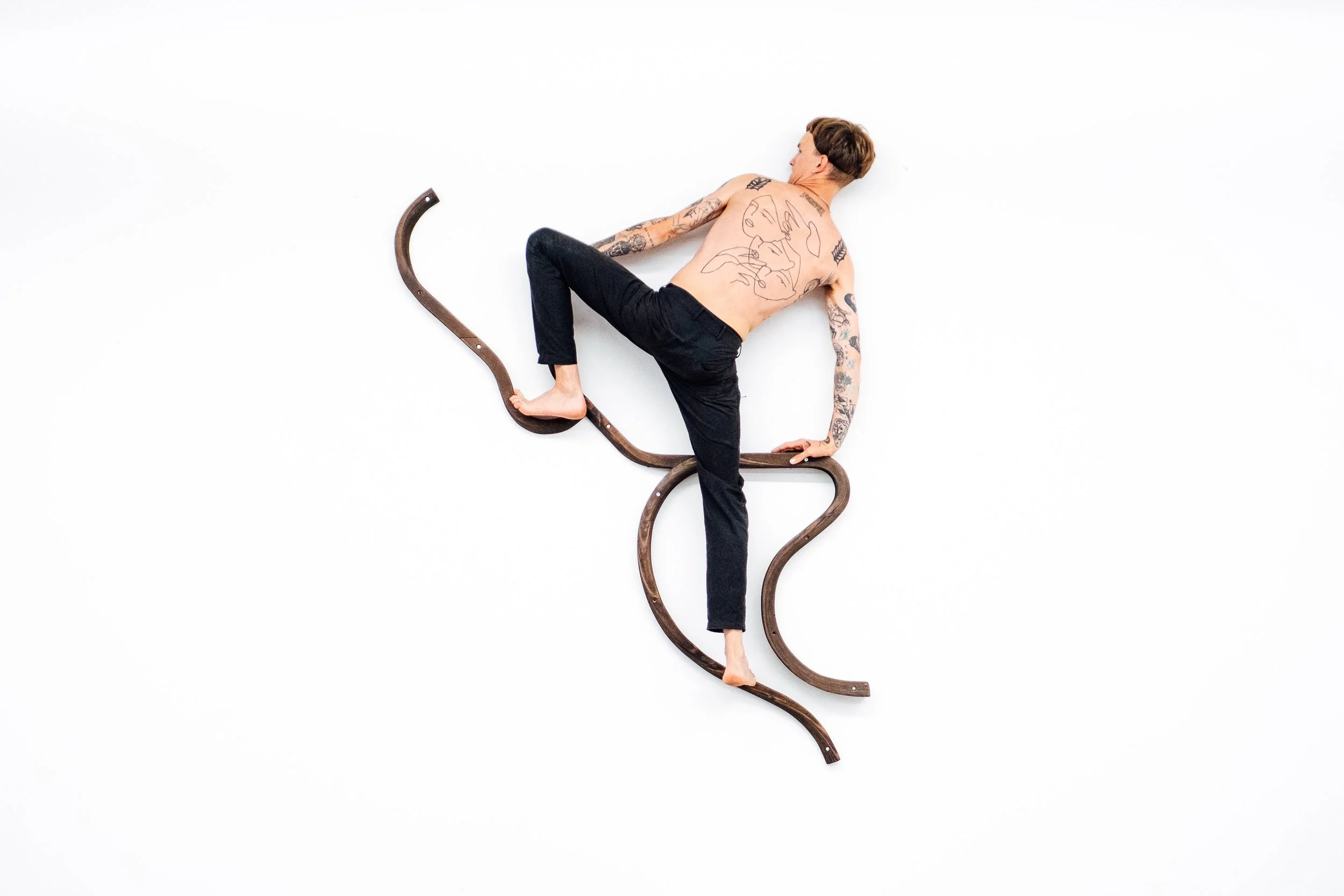 Un homme nu avec des tatouages, en équilibre sur une structure en forme de spirale en bois, sur fond blanc.