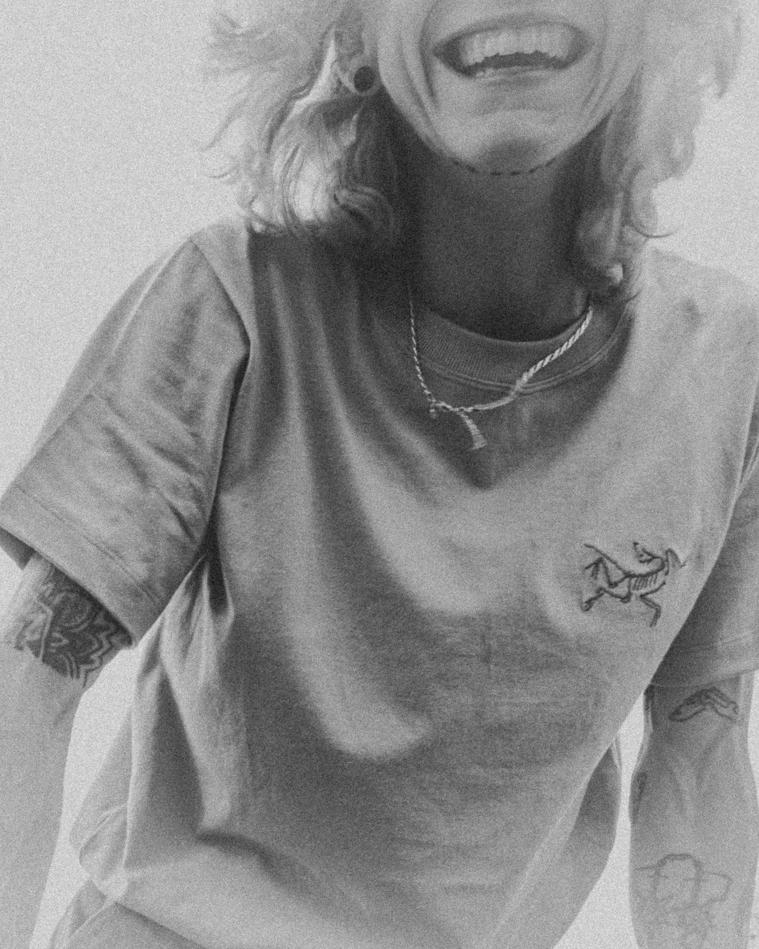 Un portrait en noir et blanc d'une personne souriante portant un t-shirt avec un tatouage sur le bras, une chaîne et une boucle d'oreille. La photo est prise de près, mettant en valeur le sourire et les détails du visage et de la peau.