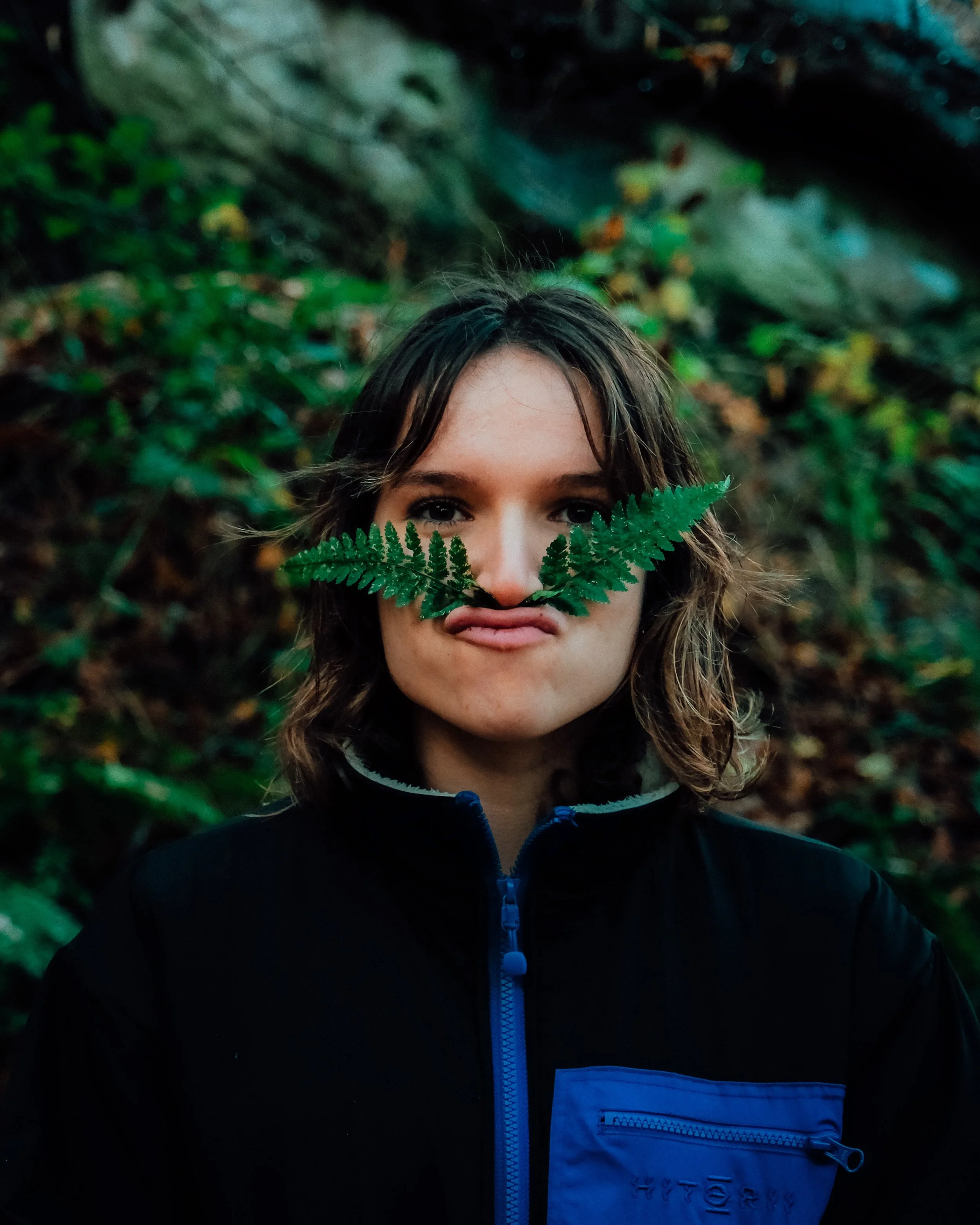 Une personne, avec des cheveux bruns, tien un feuillage vert devant son visage, dans un décor naturel avec des feuilles et de la végétation.