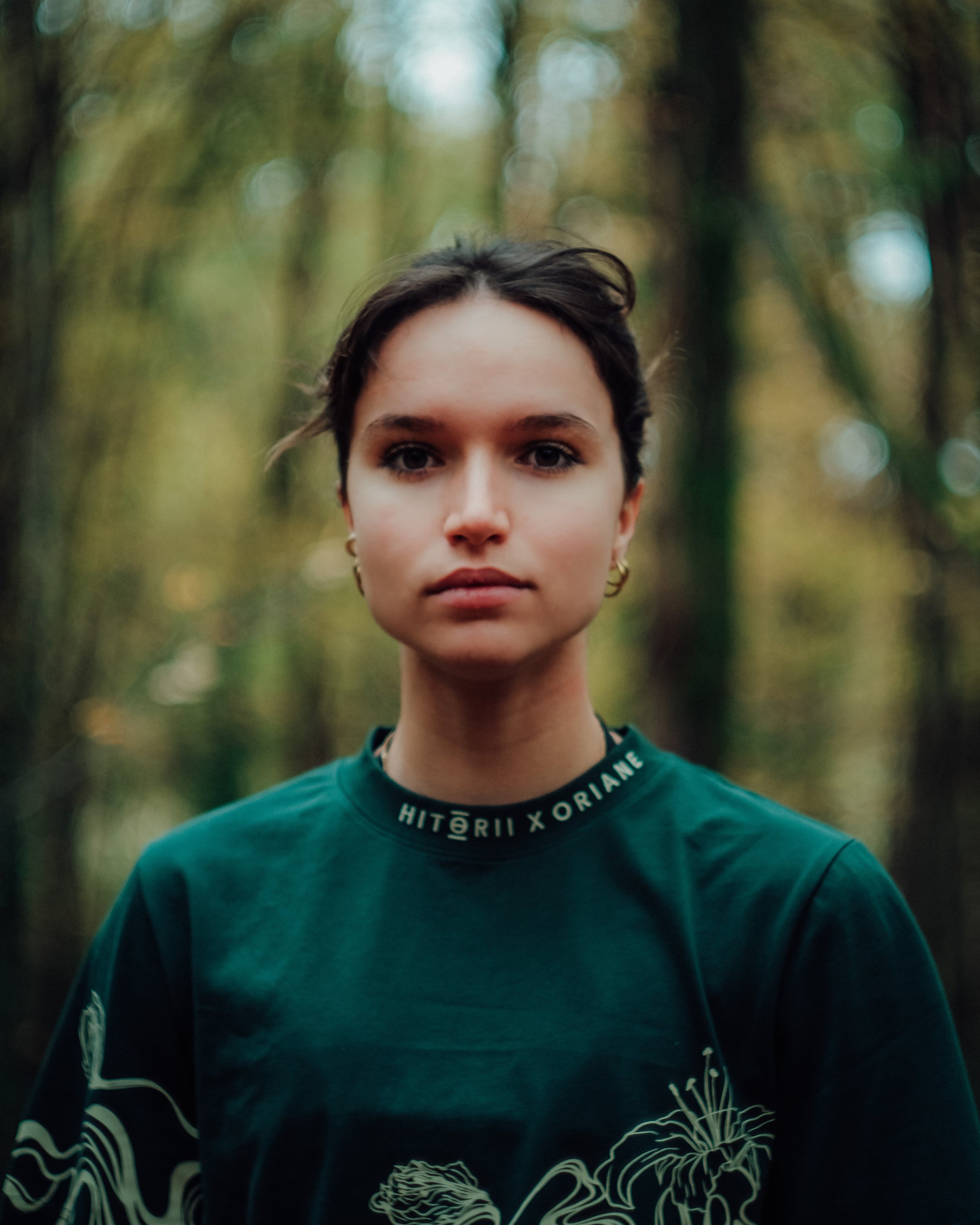 Portrait en gros plan d'une jeune femme avec un fond flou de forêt. Elle porte un t-shirt vert foncé avec un dessin floral blanc et une inscription en anglais autour du col.