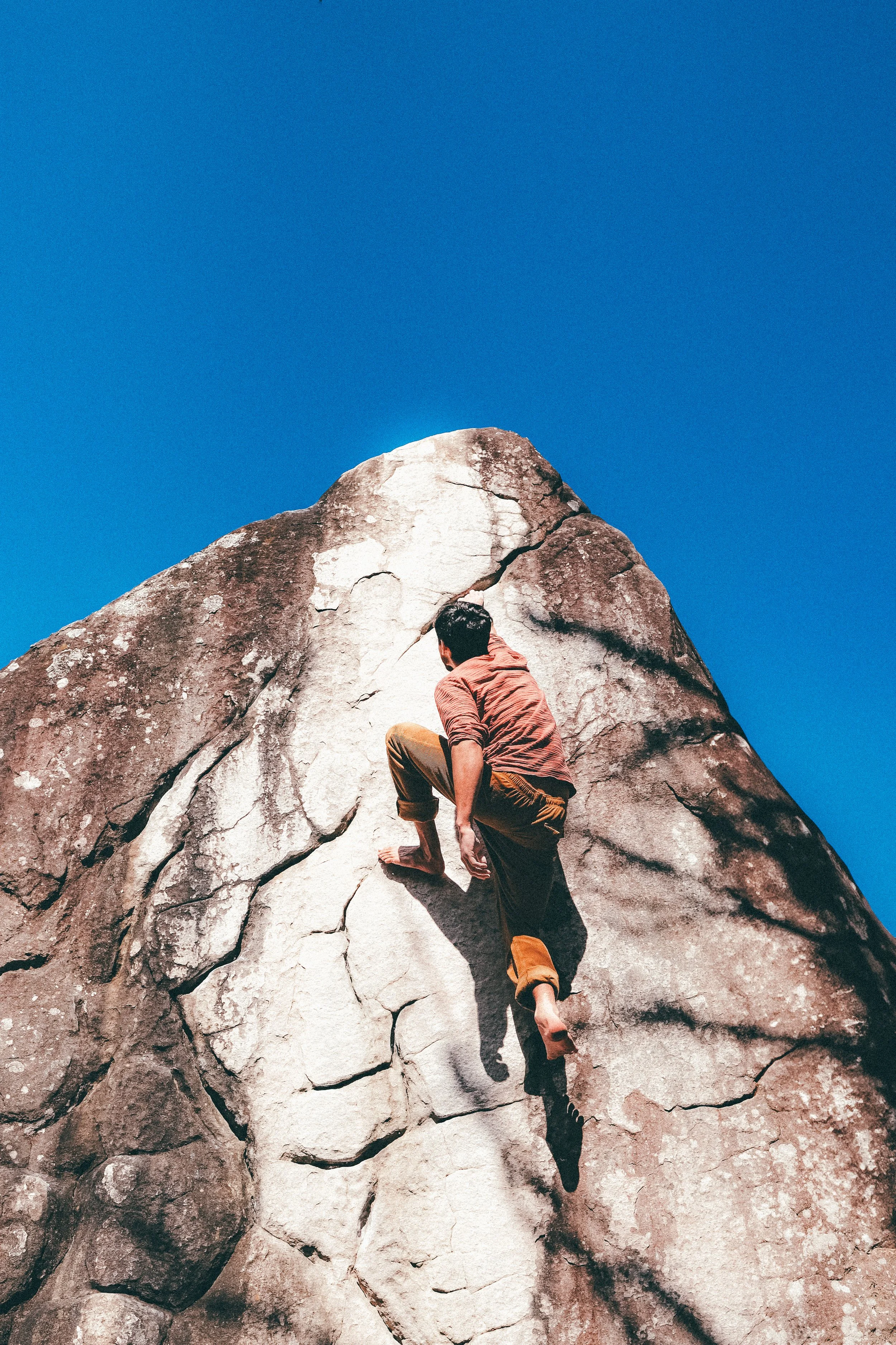 Un homme grimpe une grosse roche en plein air sous un ciel bleu clair.